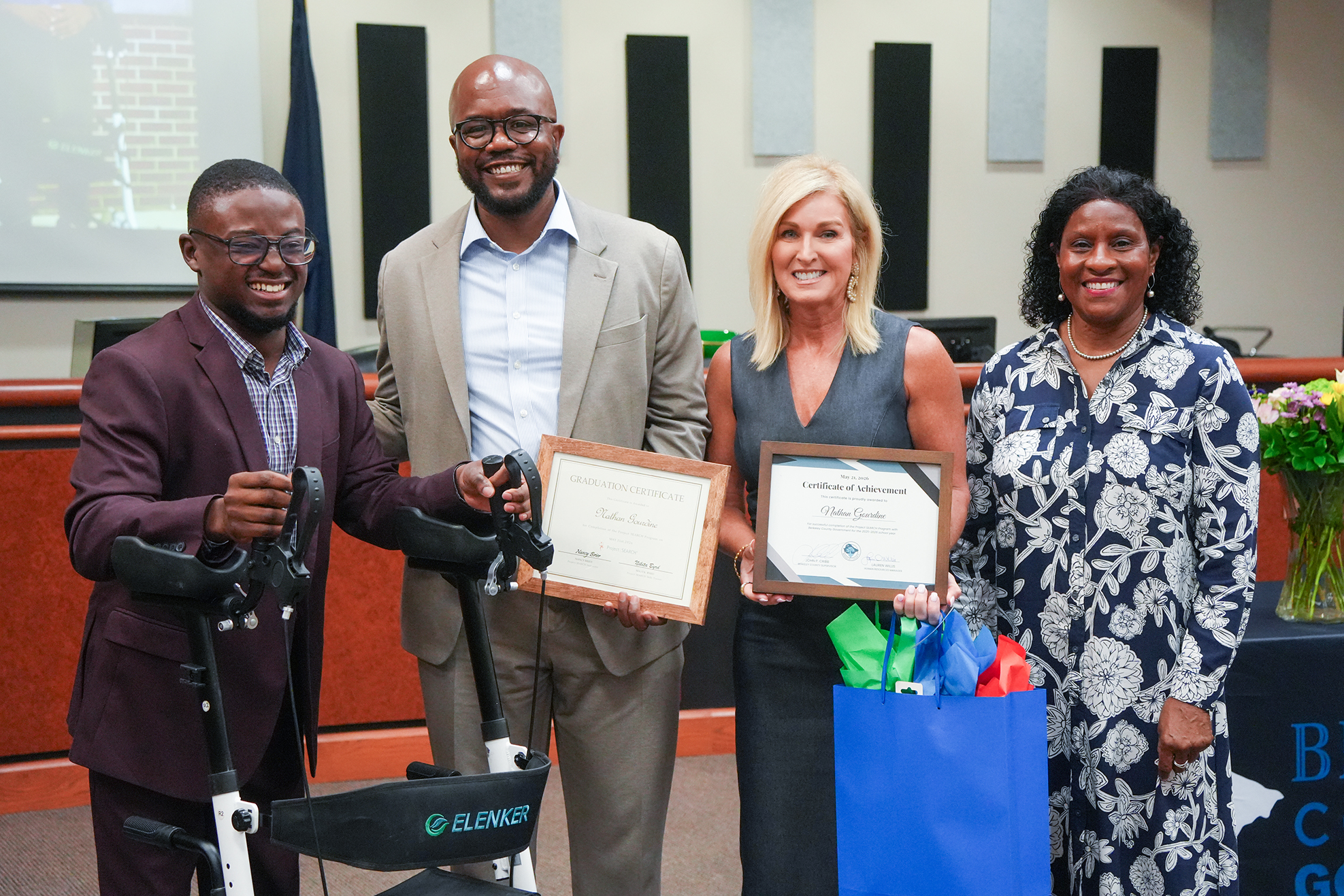 Four people stand together, two men and two women, holding certificates and a blue bag. One man holds a walker.