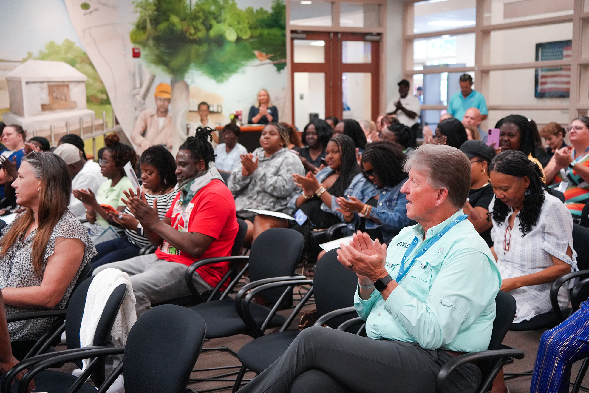 Group of people seated in chairs clapping, one man wearing blue shirt, seated in front row.