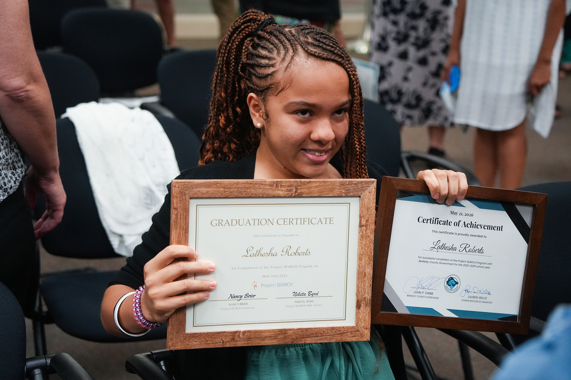 A young girl in a green dress holds two framed certificates in front of an audience.