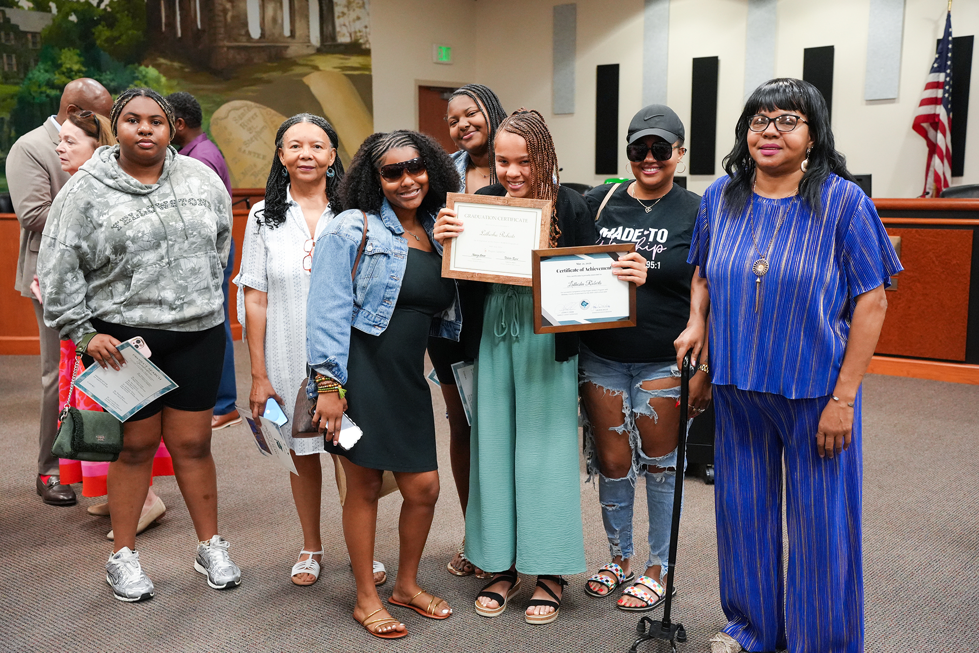 A group of women, some holding certificates, pose in a room with a wall featuring a mural and an American flag.