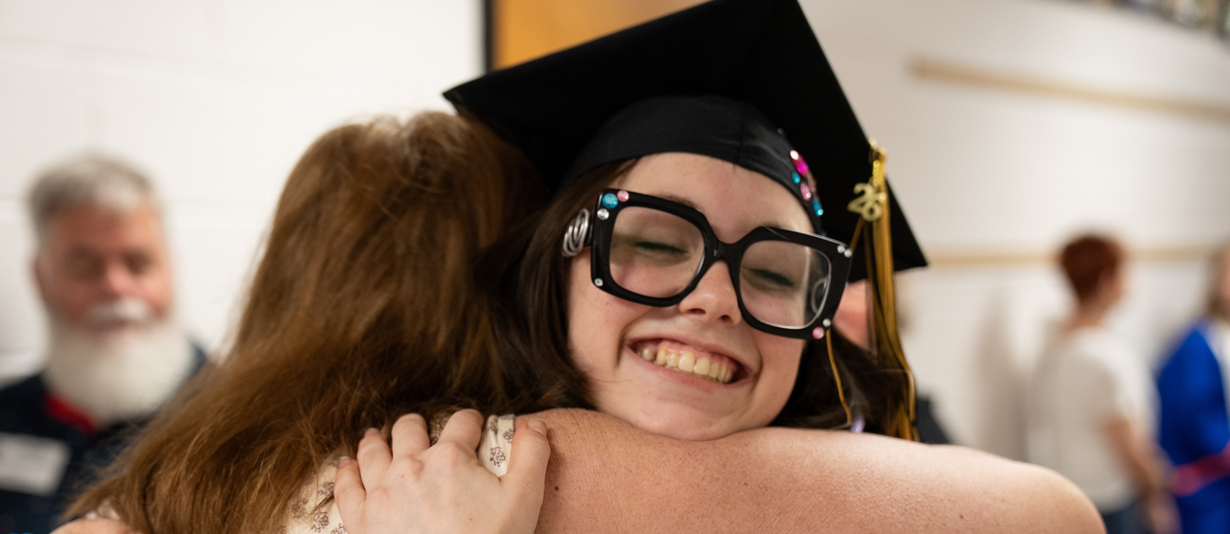 A woman in a black cap and gown hugs another woman in a white dress, smiling. Two other people are in the background.