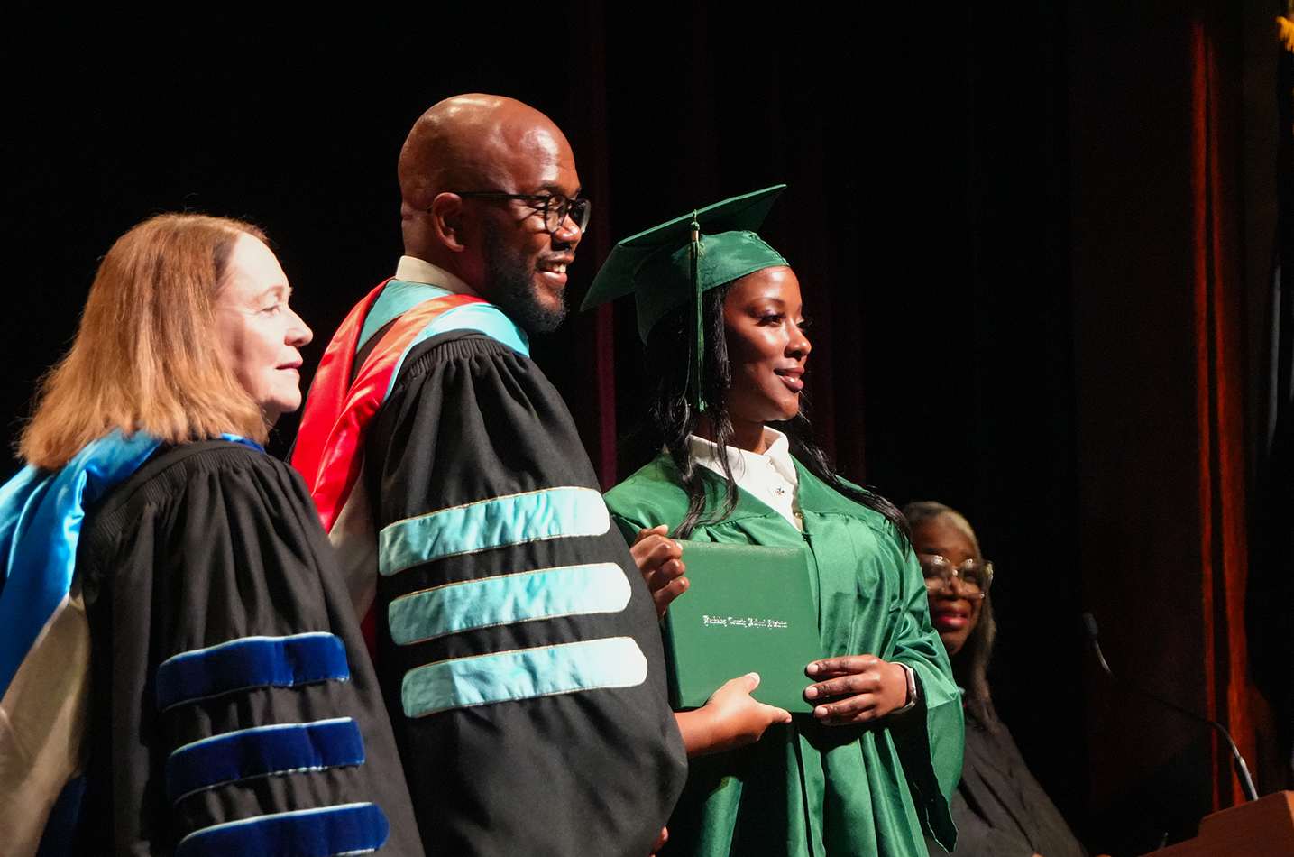 Graduates in academic robes, one holding a diploma, stand on stage with a faculty member in the background.