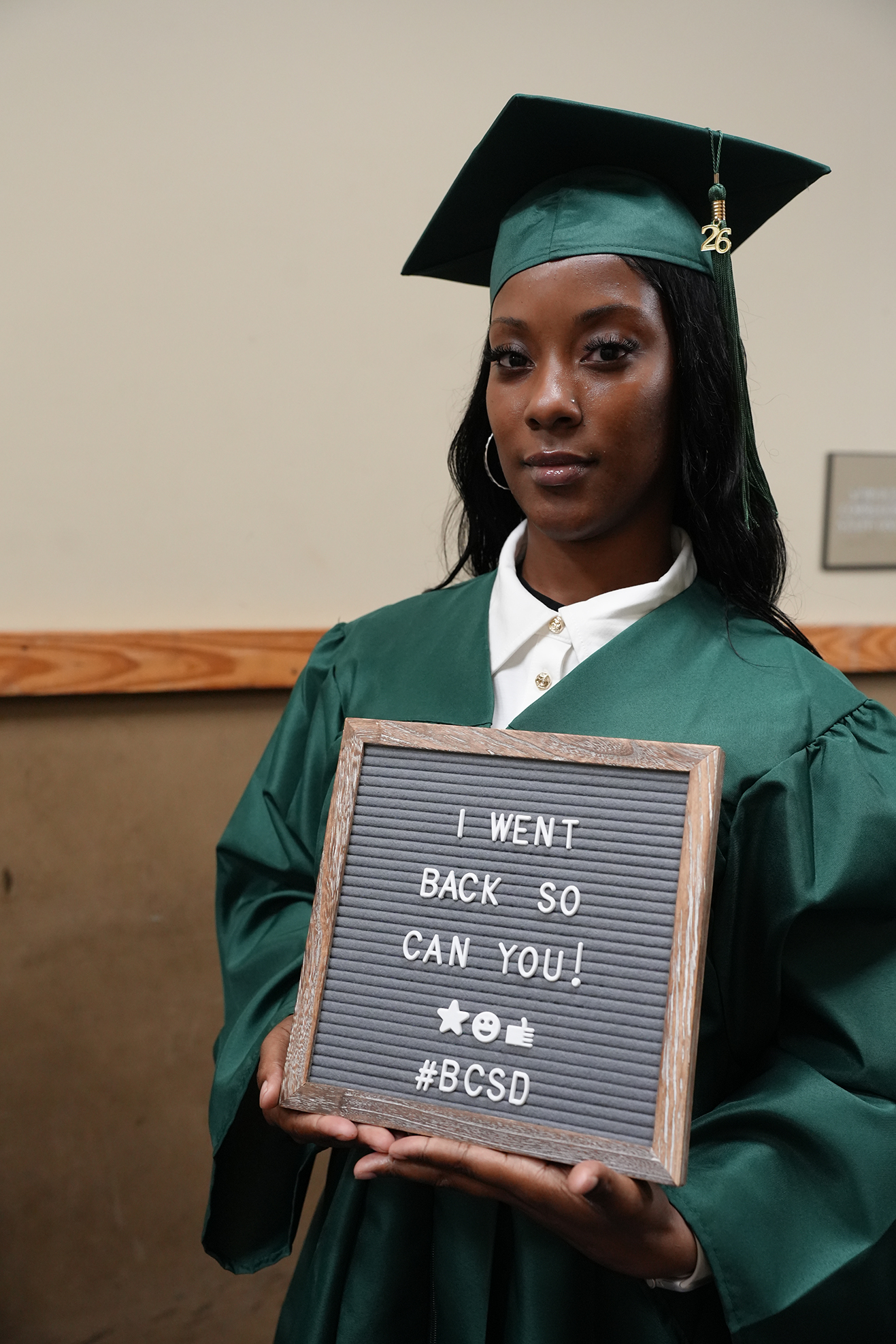 A woman in a green graduation gown and cap holds a sign that reads "I went back so you can you!"