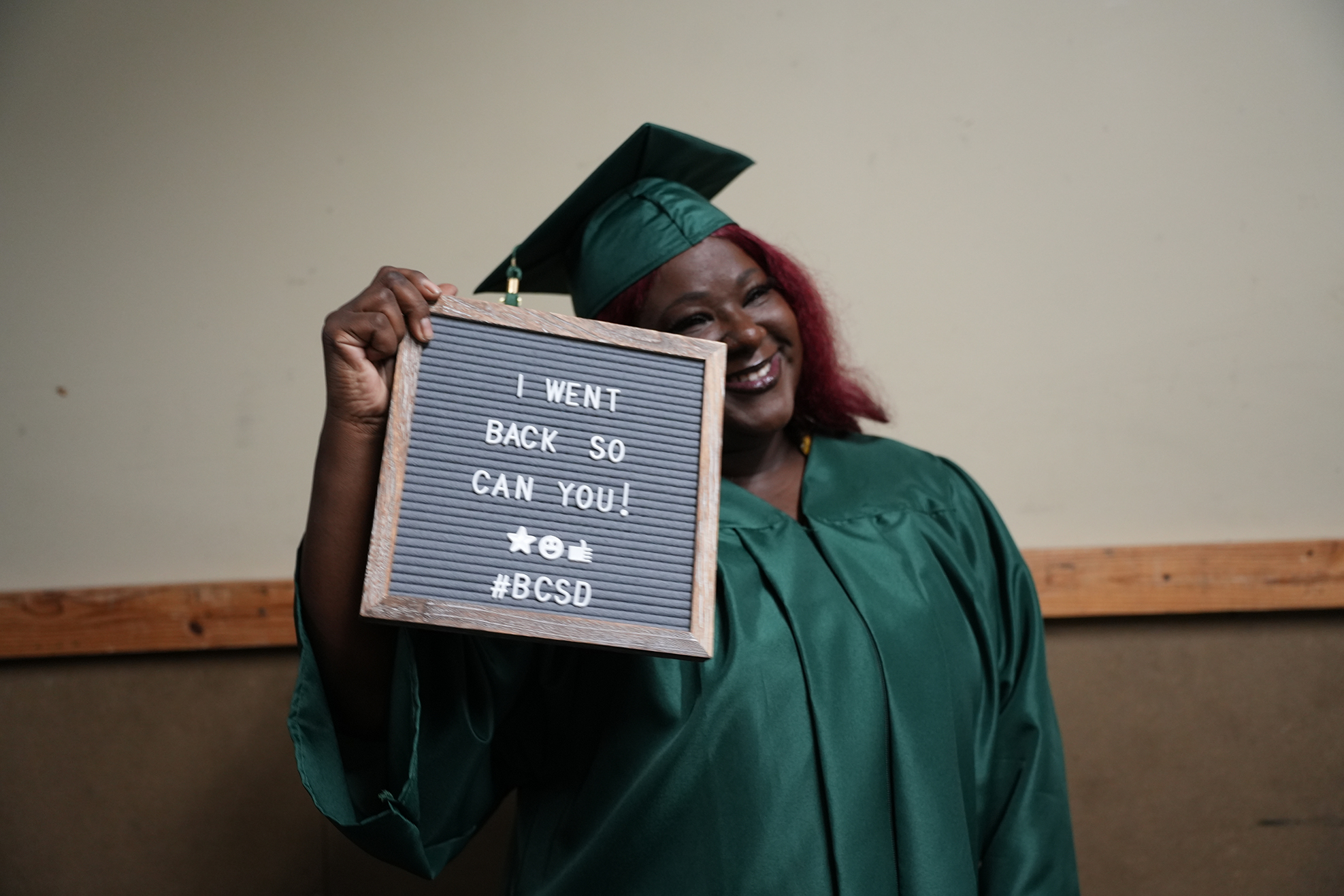 Person in a green graduation gown and cap holds a sign with white writing on a blackboard.