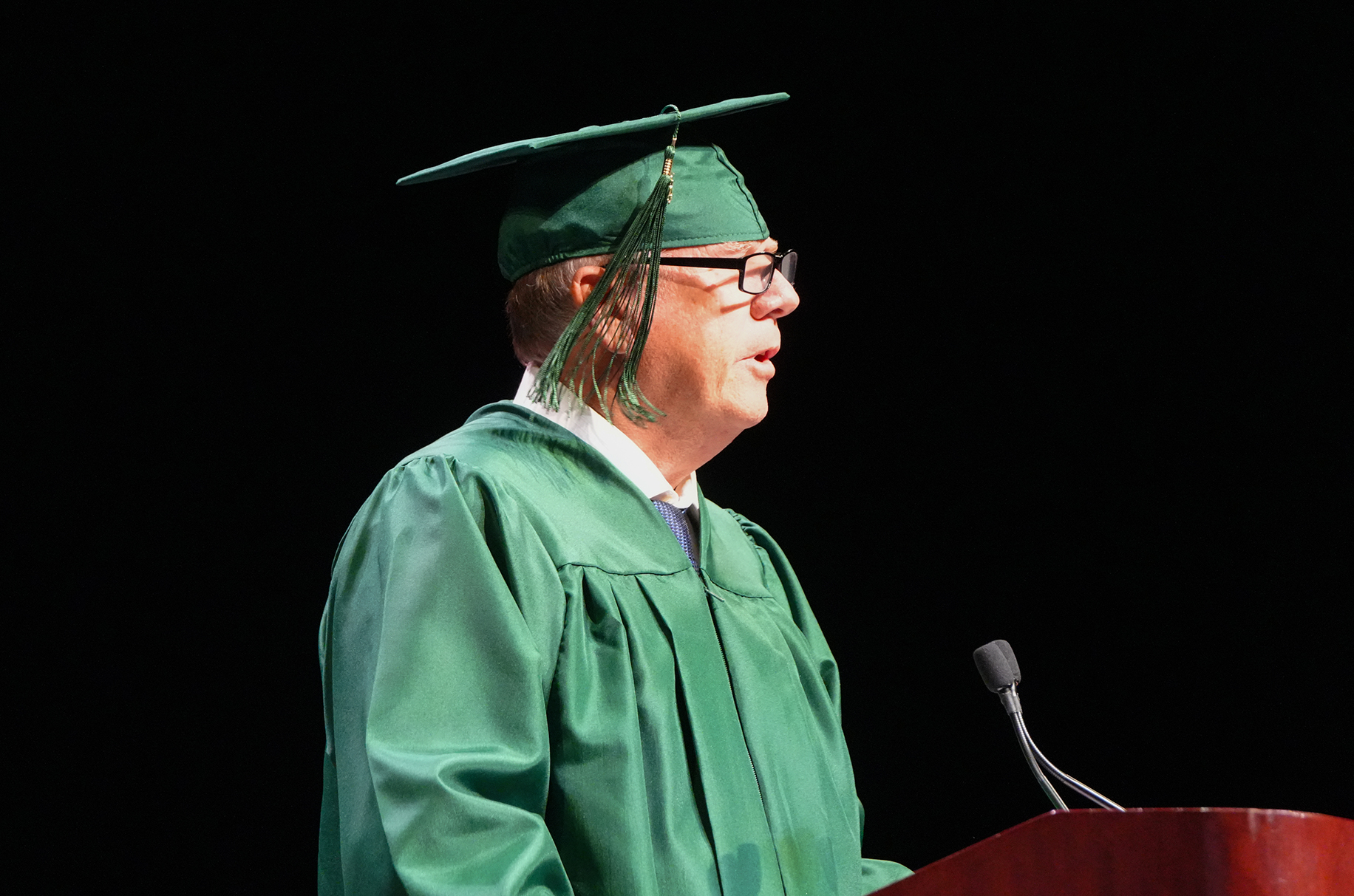 A person in a green graduation gown and cap stands at a podium, speaking into a microphone.