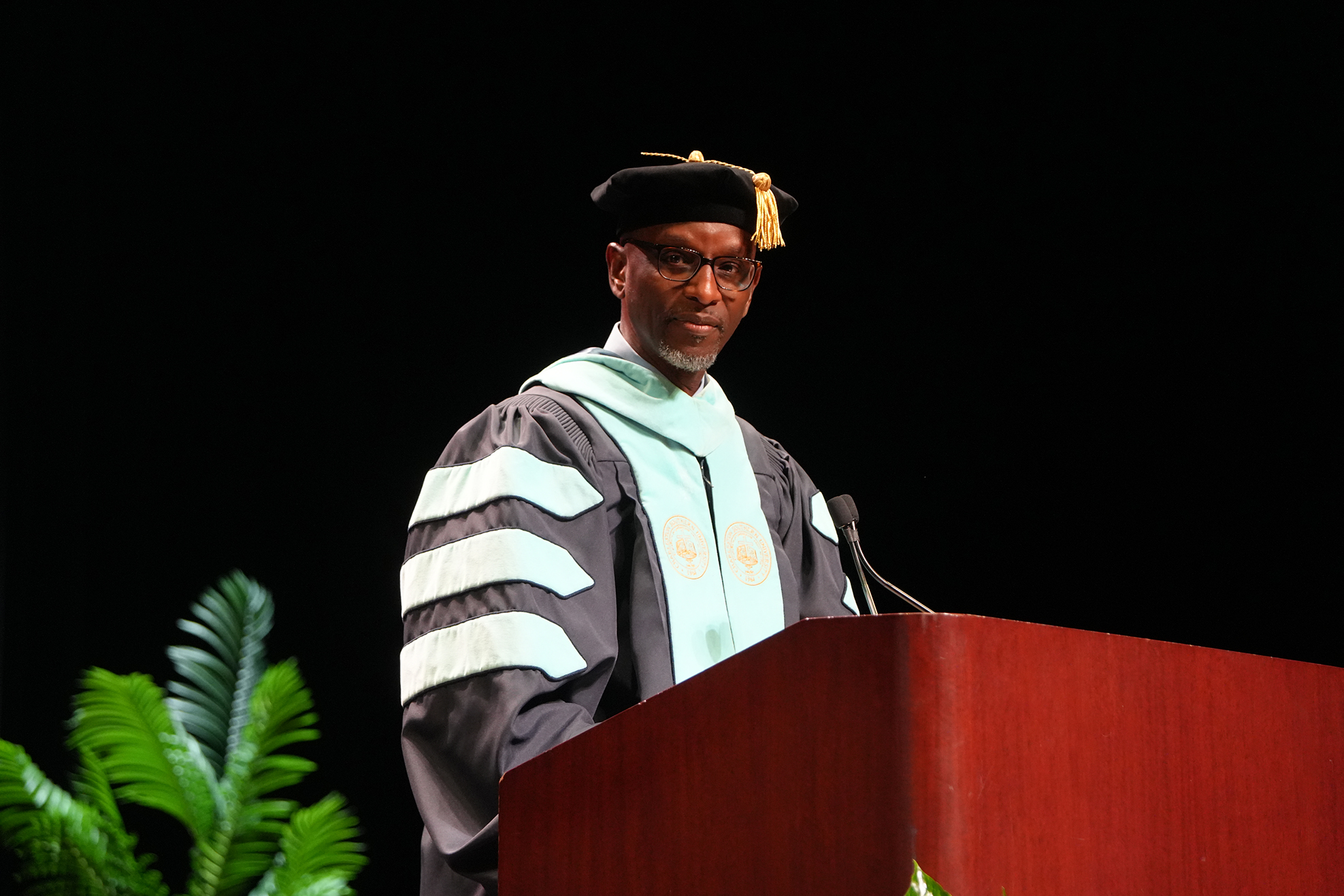 Man in a black academic gown and hat stands at a podium with a microphone.