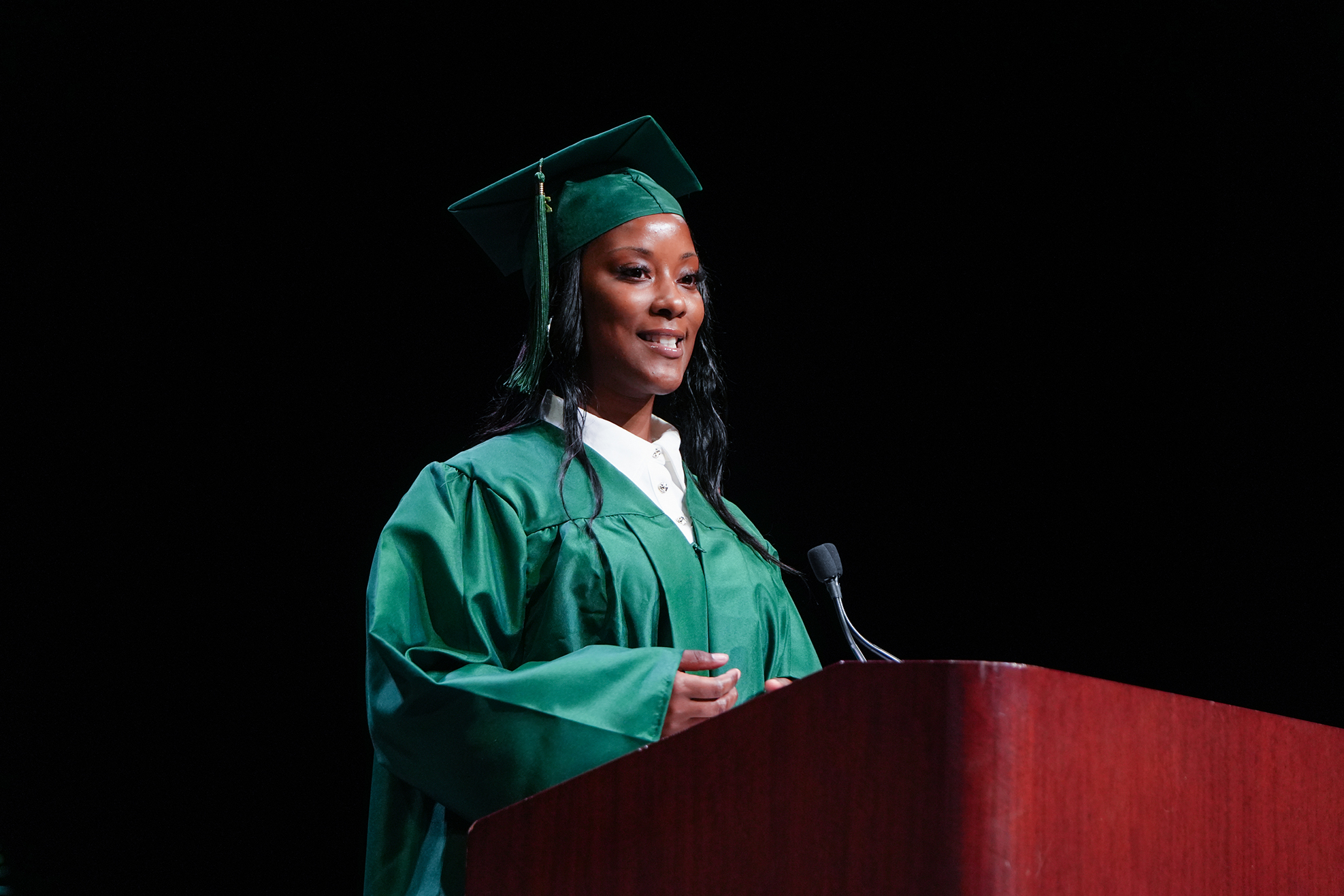 Woman in green graduation cap and gown, smiling, stands at a podium with microphone.