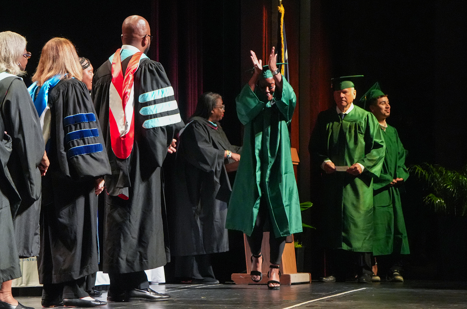 Several people in green gowns on a stage, one raising an arm. Faculty in black gowns stand nearby.