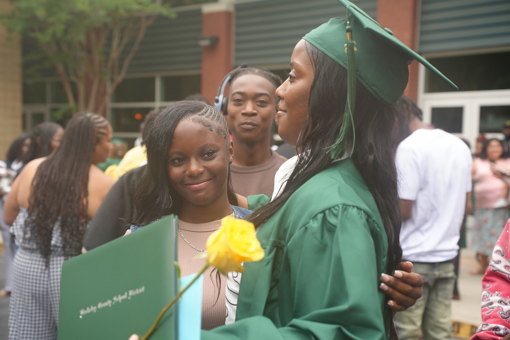 A woman in a green gown, holding a diploma, shares a moment with a friend. They smile.