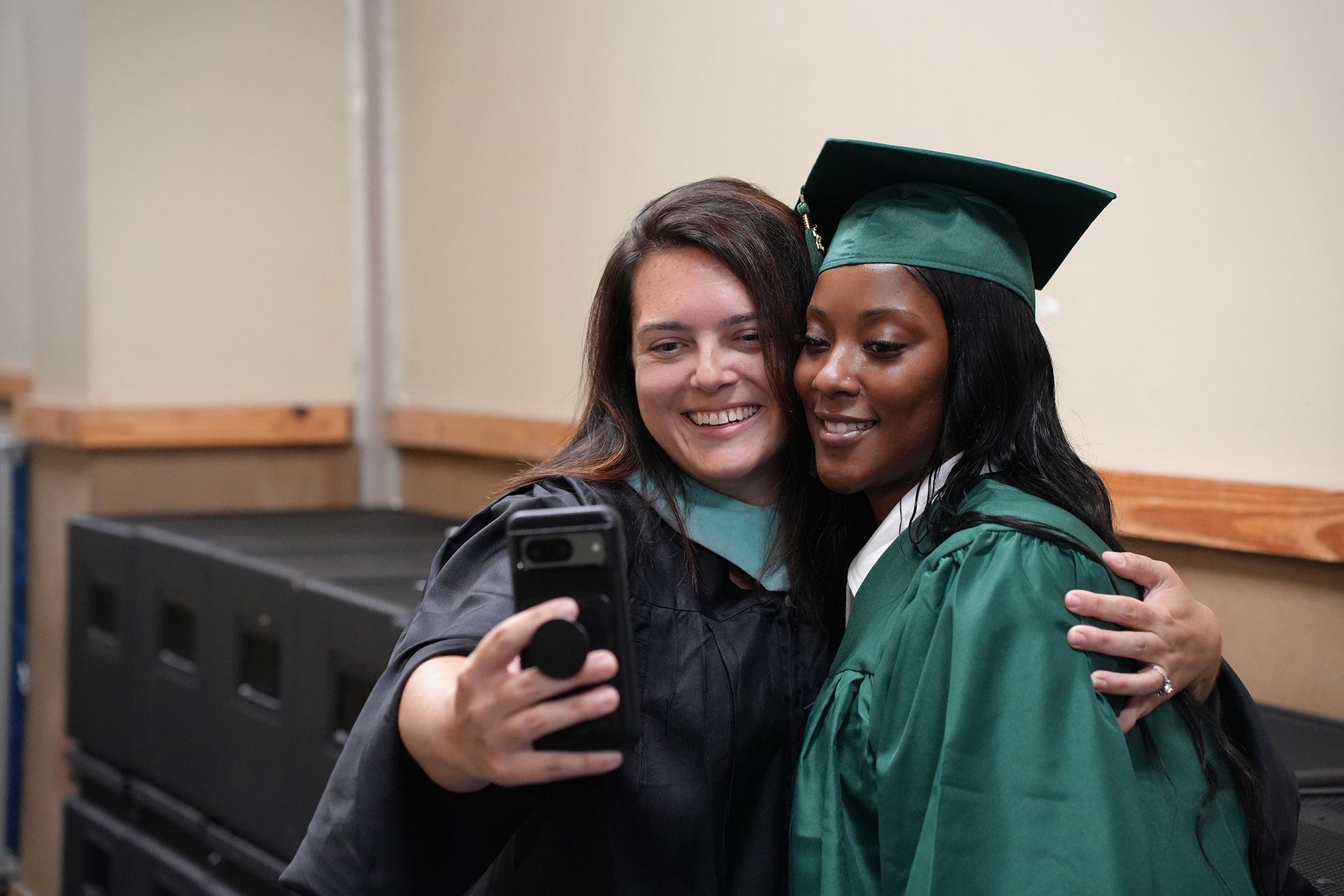 Two women in graduation gowns, one taking a selfie and the other smiling, pose together.