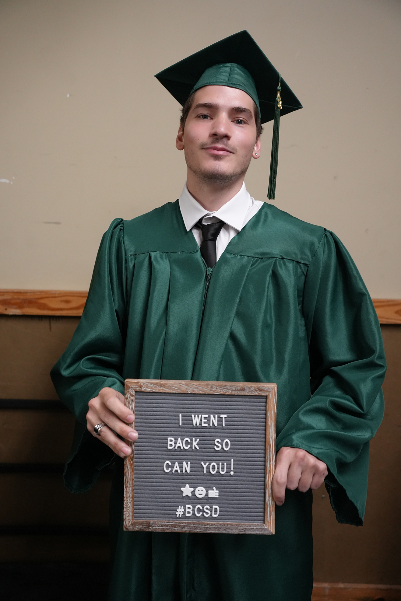 Man in green graduation gown holding sign that reads "I WENT BACK SO CAN YOU! #BCSd".
