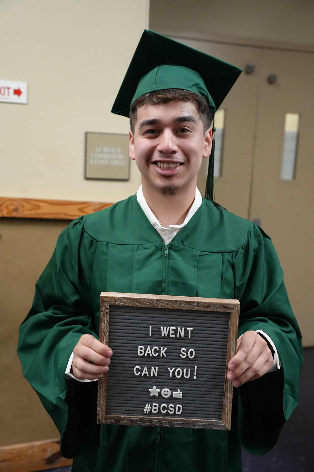 A person in a green graduation gown holds a sign reading "I WENT BACK SO CAN YOU! #BCSGrad #BCSGrad2024."