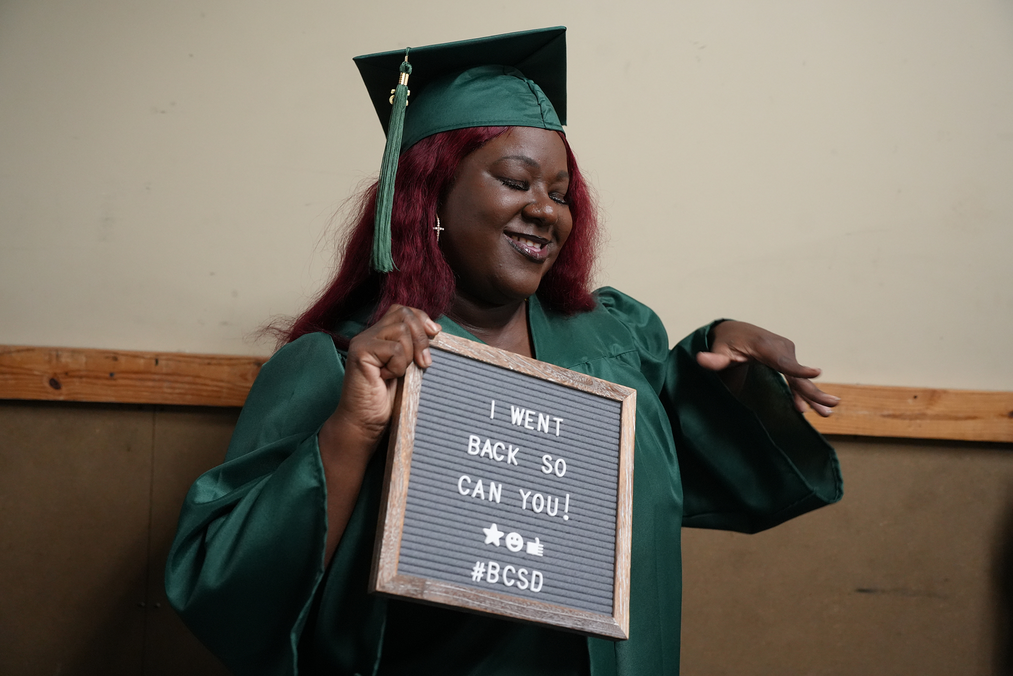Person in green graduation gown holds a sign that says "I WENT BACK SO I CAN BE YOU."