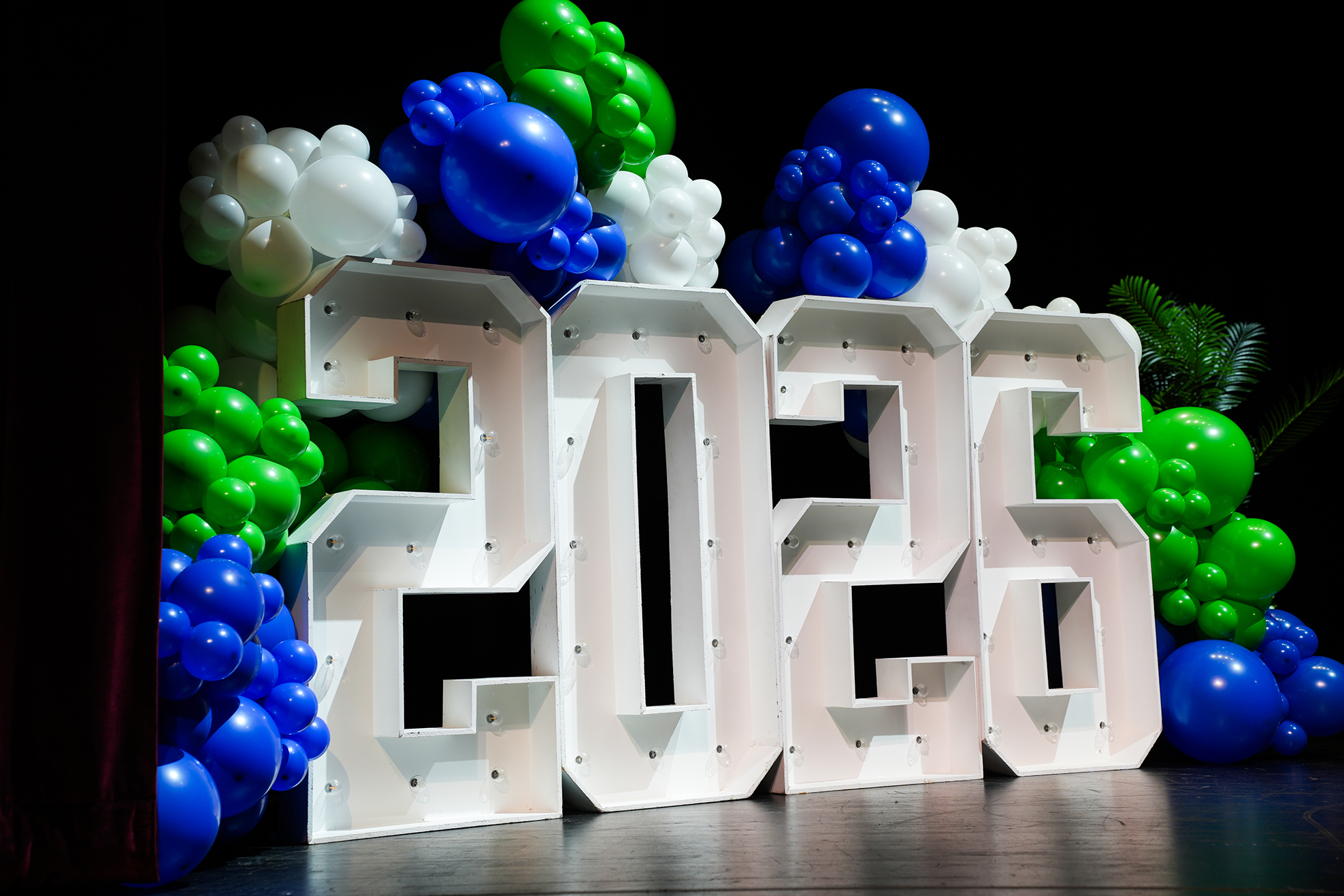 A white 2023 sign with letters in front of blue, white, and green balloons on a black background.