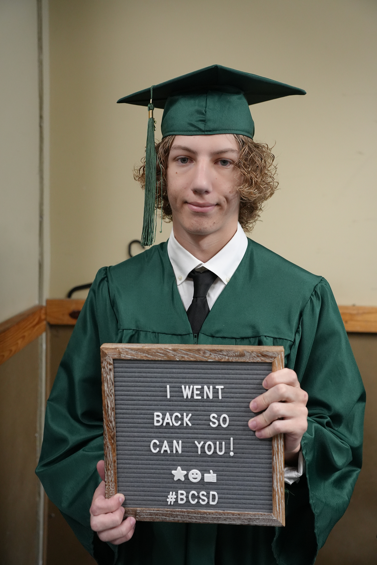 Man in green graduation gown and cap holds sign that reads "I WENT BACK SO CAN YOU!" and hashtag BCSD.