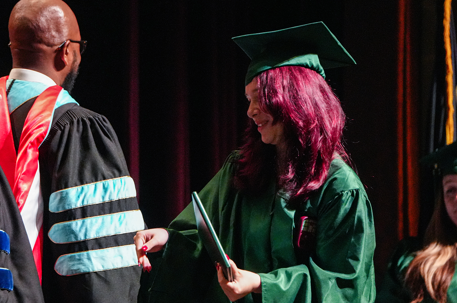 A woman in green graduation attire hands a diploma to a man in black and red academic dress.