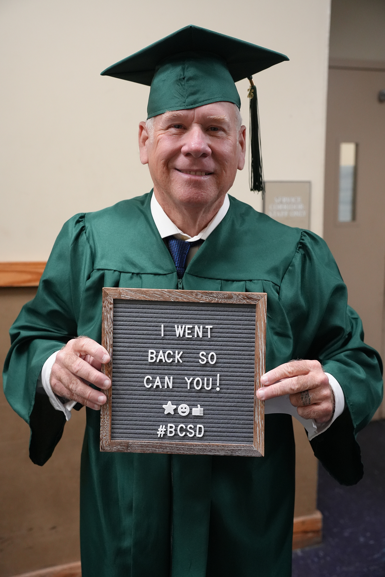 Man in a green graduation gown holding a sign that says "I WENT BACK SO CAN YOU!" Indoors.