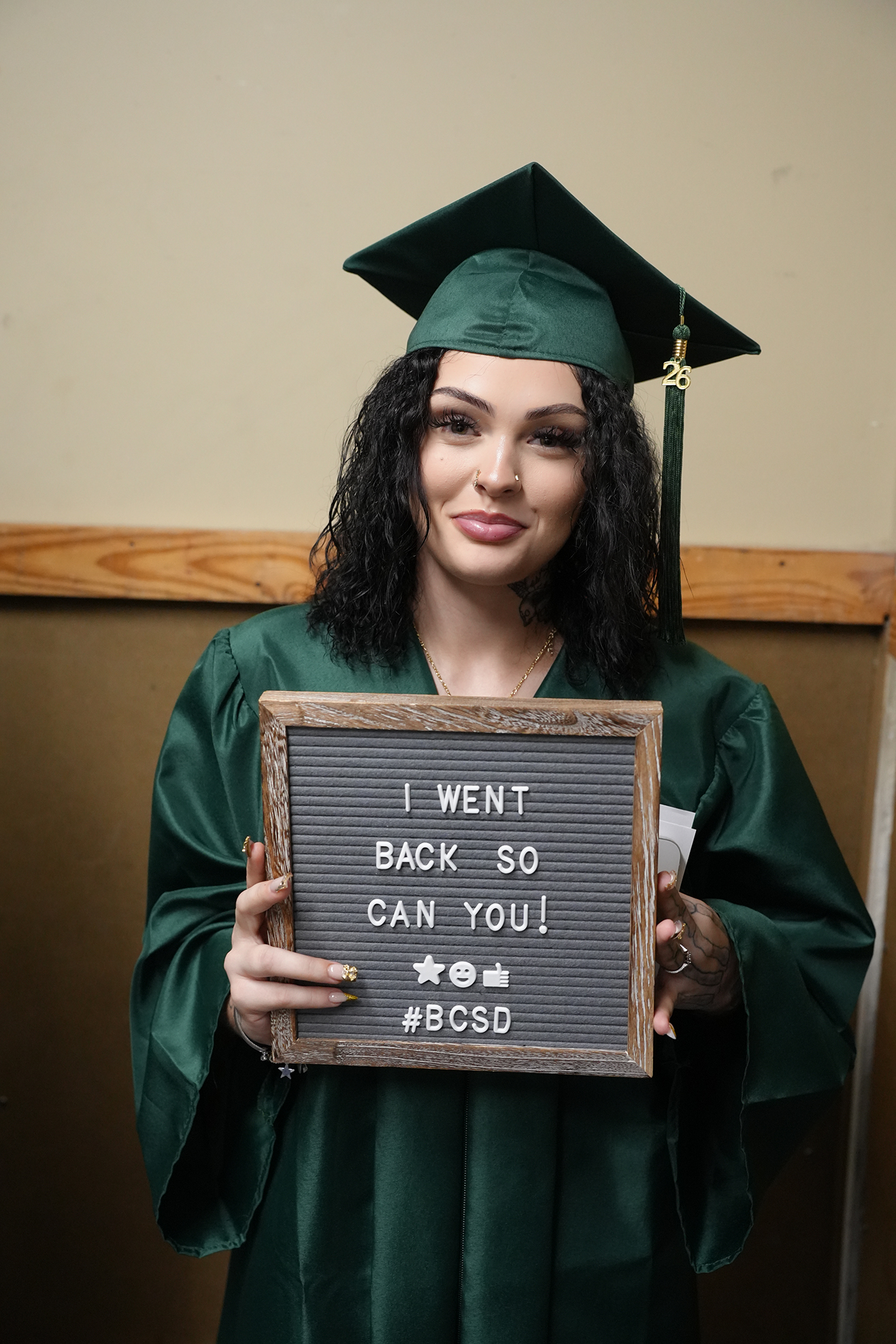 Person in green graduation cap and gown holds sign that reads "I WENT BACK SO YOU CAN TOO! #BCSGrad".