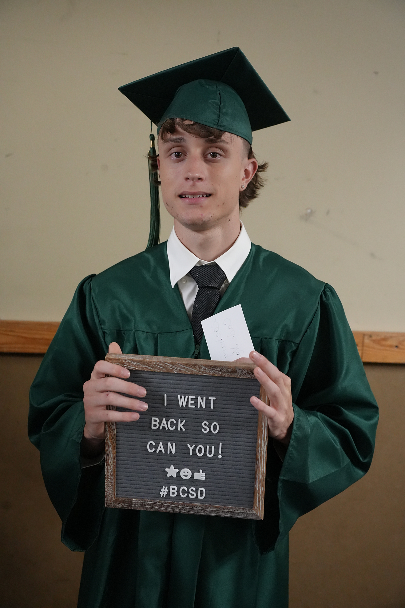 A person in a green graduation gown holding a sign that reads "I WENT BACK SO YOU CAN YOU! #BCSD".