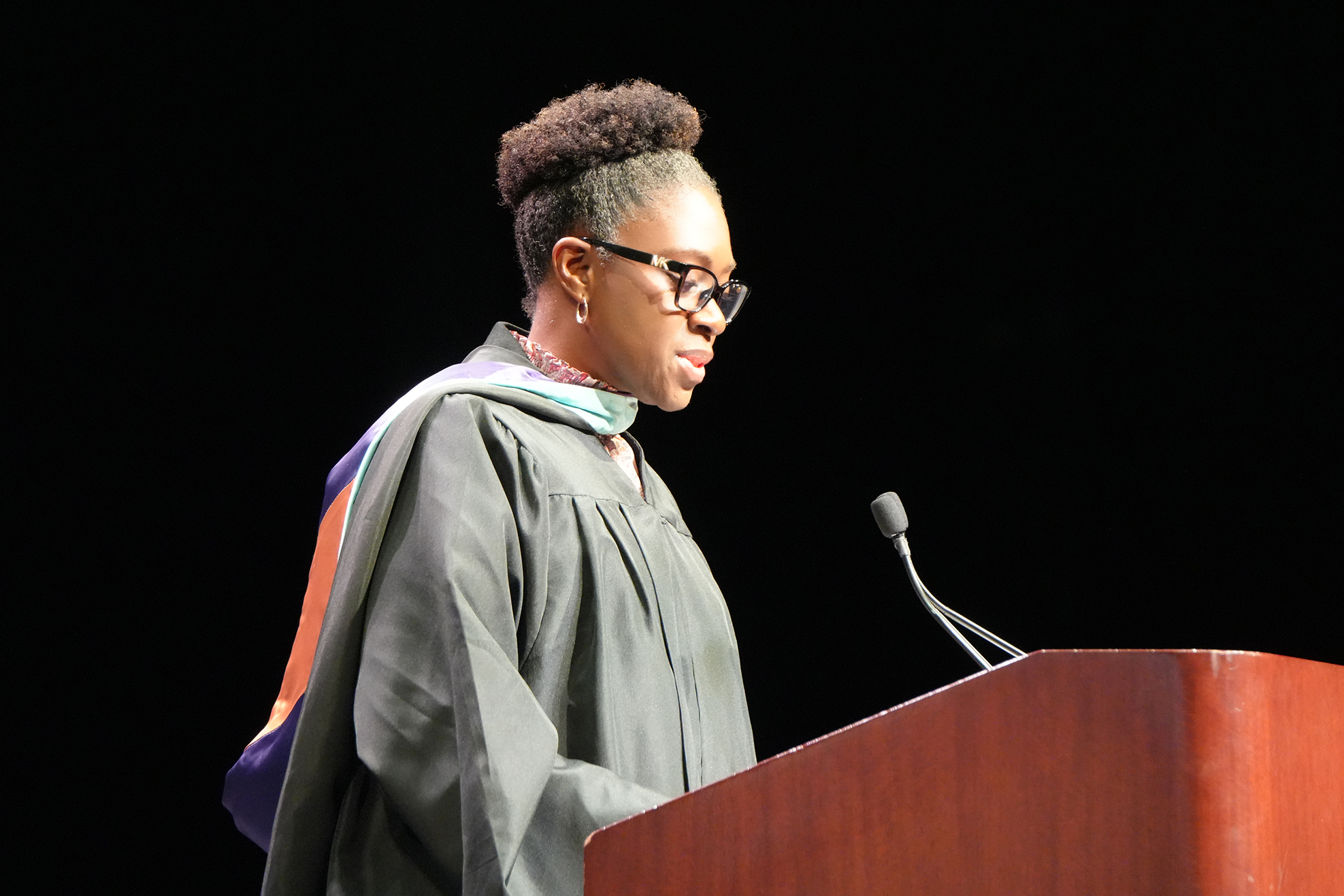 A person in a graduation gown stands at a podium with a microphone, wearing glasses.