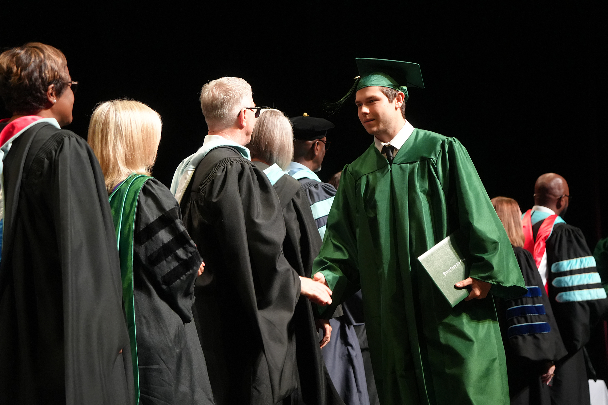Graduates in caps and gowns at a ceremony, one in green holding a book, others in black.