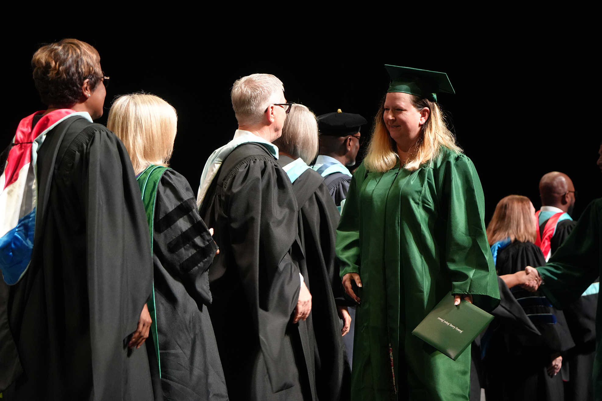 Graduates in academic attire stand in line; one woman in green holds a diploma. Behind them, other people in academic dress are also present.