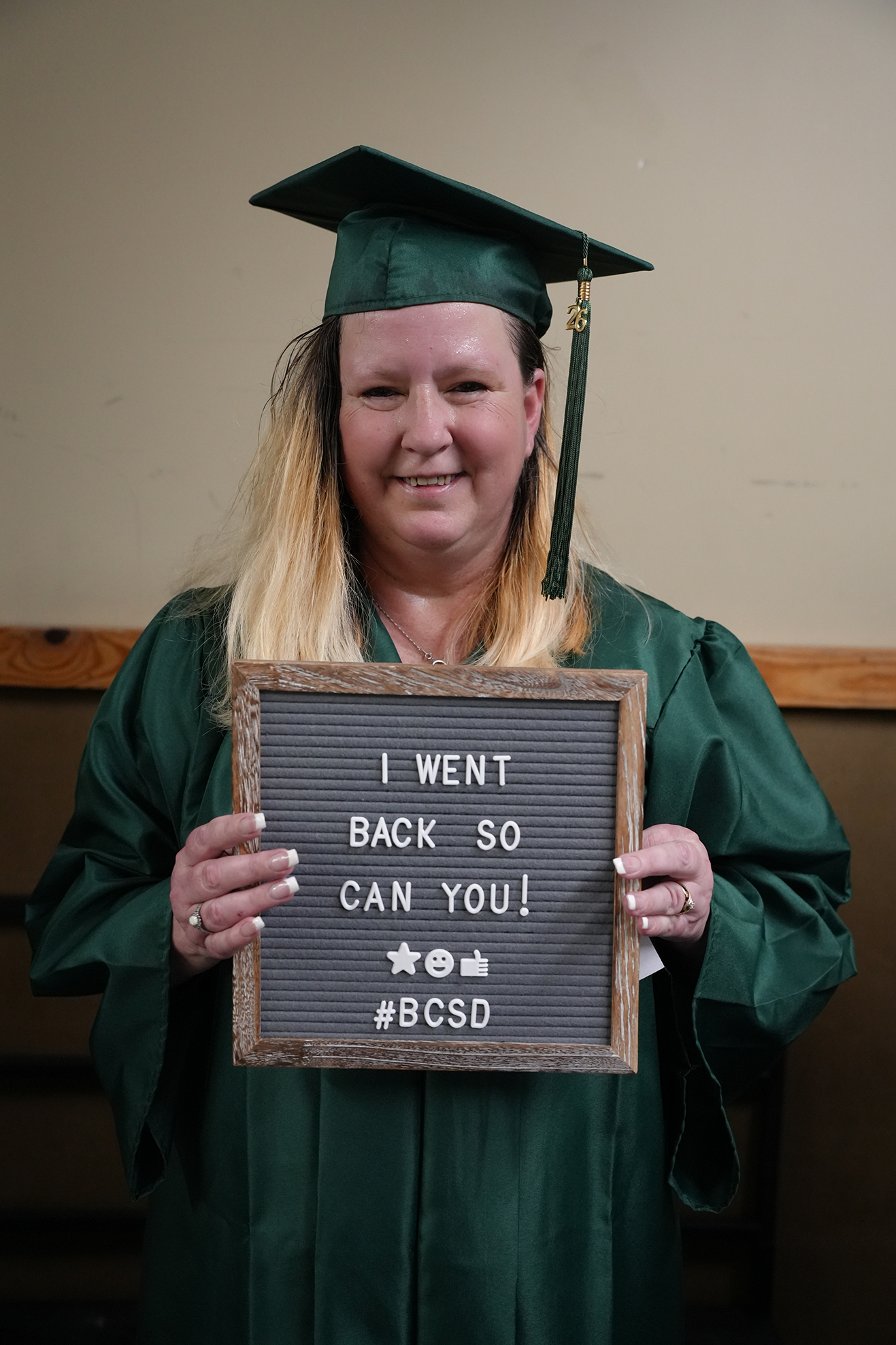 A woman in a graduation gown holds a sign that reads "I WENT BACK SO CAN YOU! #BCSD."