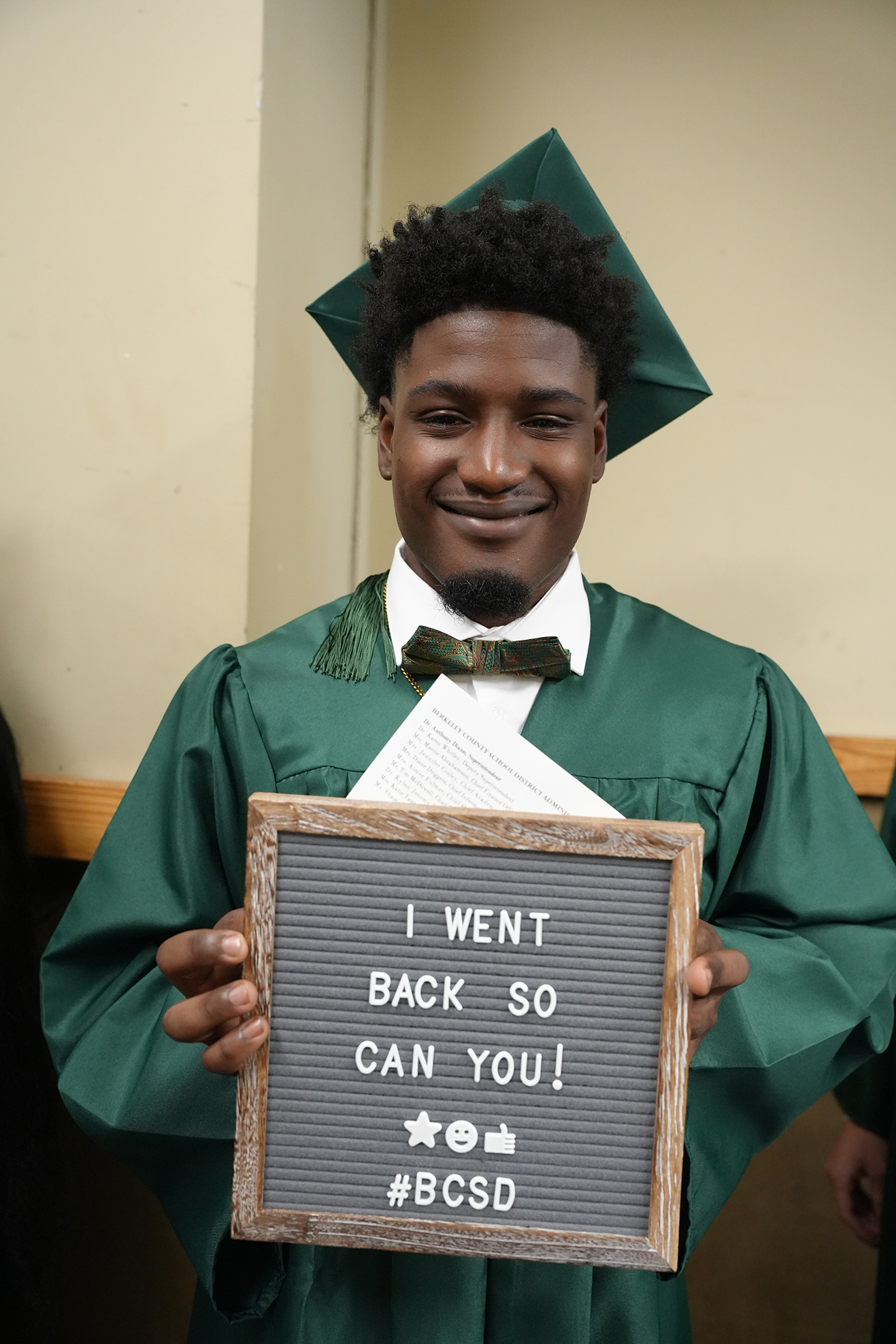 A young man in a green graduation gown holds a chalkboard sign saying "I WENT BACK SO CAN YOU!"