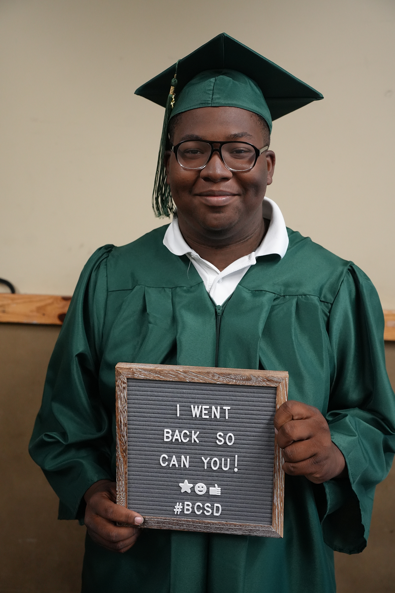 A person in a green graduation gown and cap holds a sign with a message: "I WENT BACK SO CAN YOU! #BCSd".