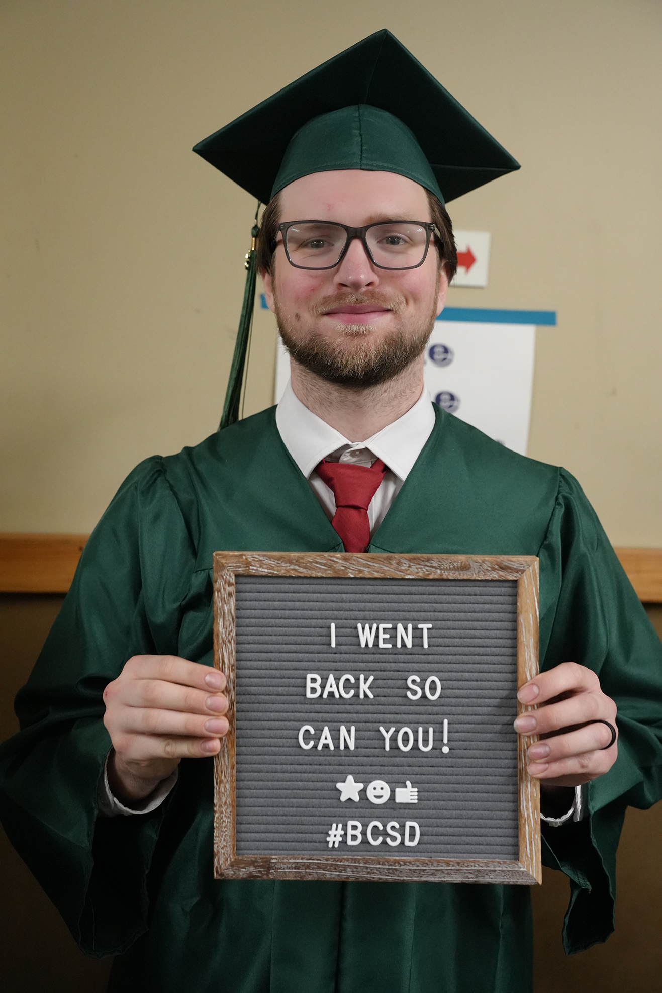Man in green graduation cap and gown holds sign that reads "I WENT BACK SO YOU CAN YOU!"