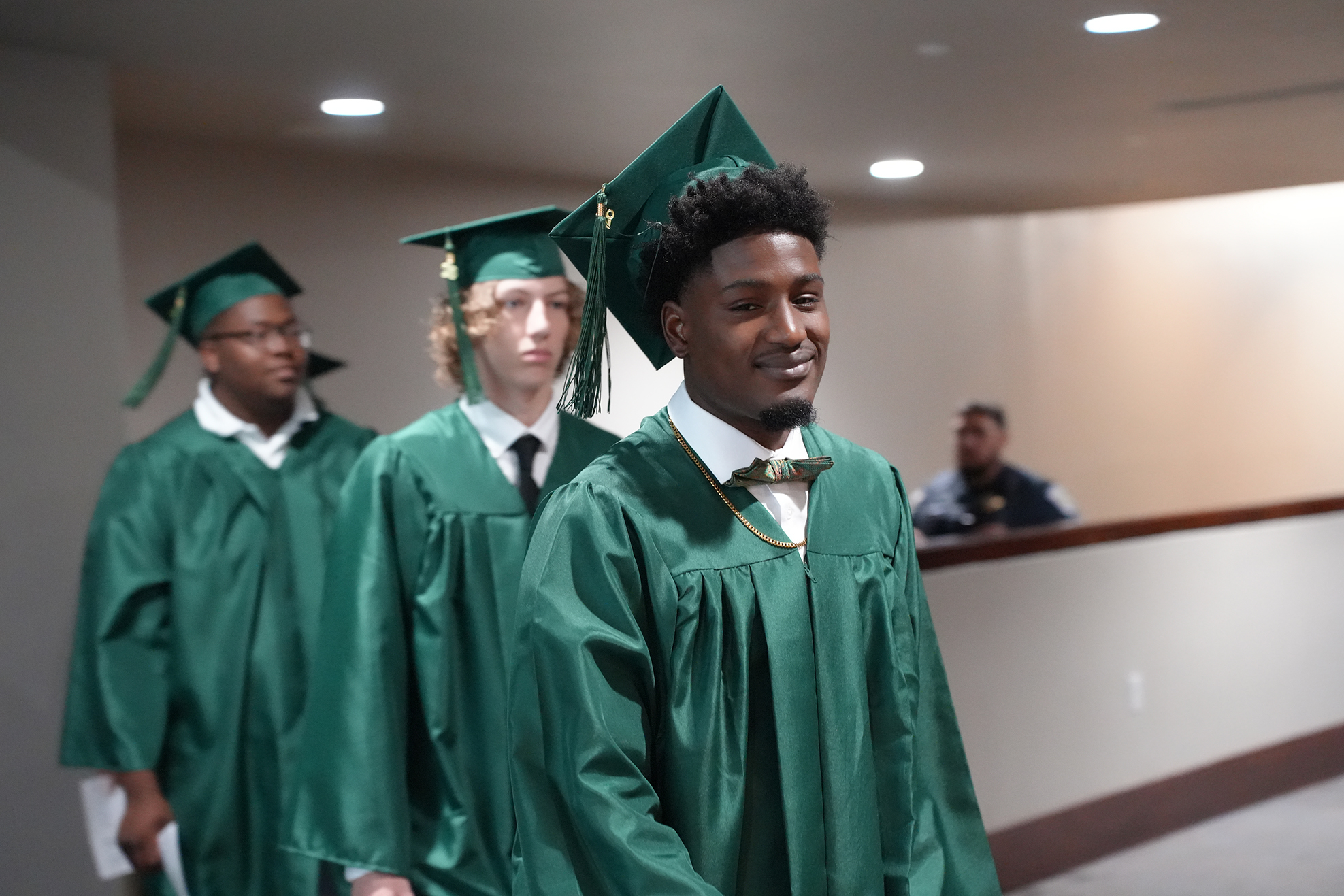 Three people in green graduation gowns and caps walk through a hallway. One looks back, smiling.