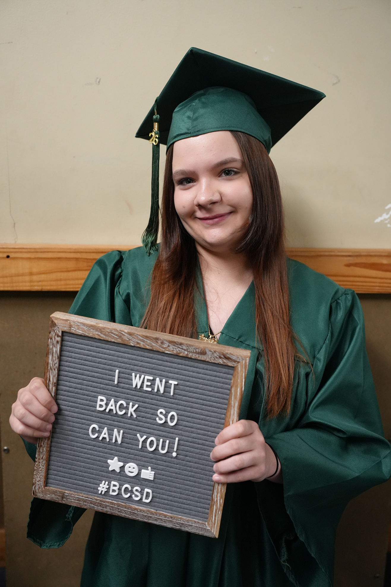 A person in green graduation attire holds a sign that says, "I WENT BACK SO CAN YOU!" Behind them is a wooden wall.