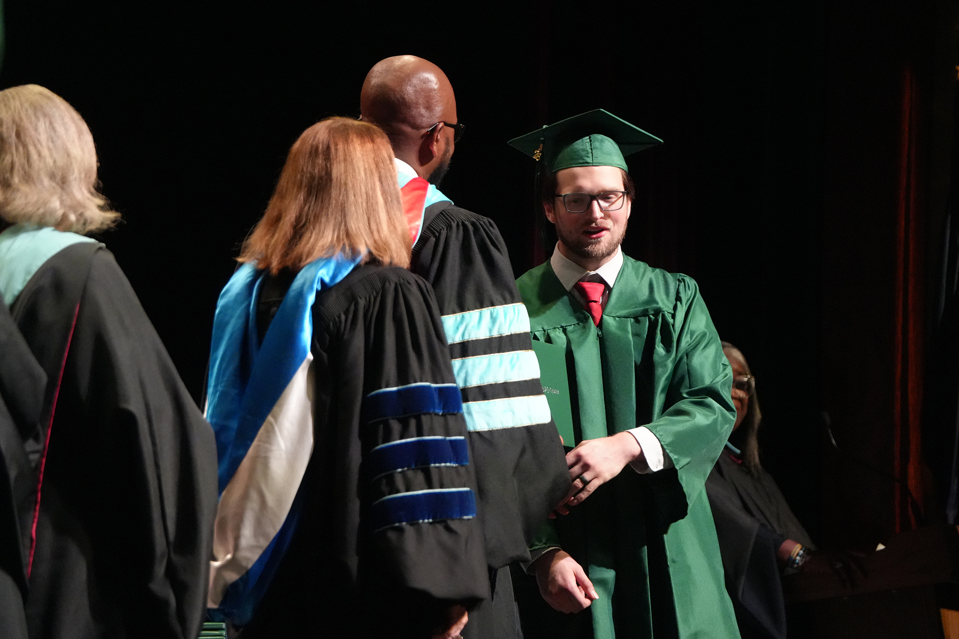 A group of individuals in academic gowns and caps, one wearing a green graduation gown, on a stage.
