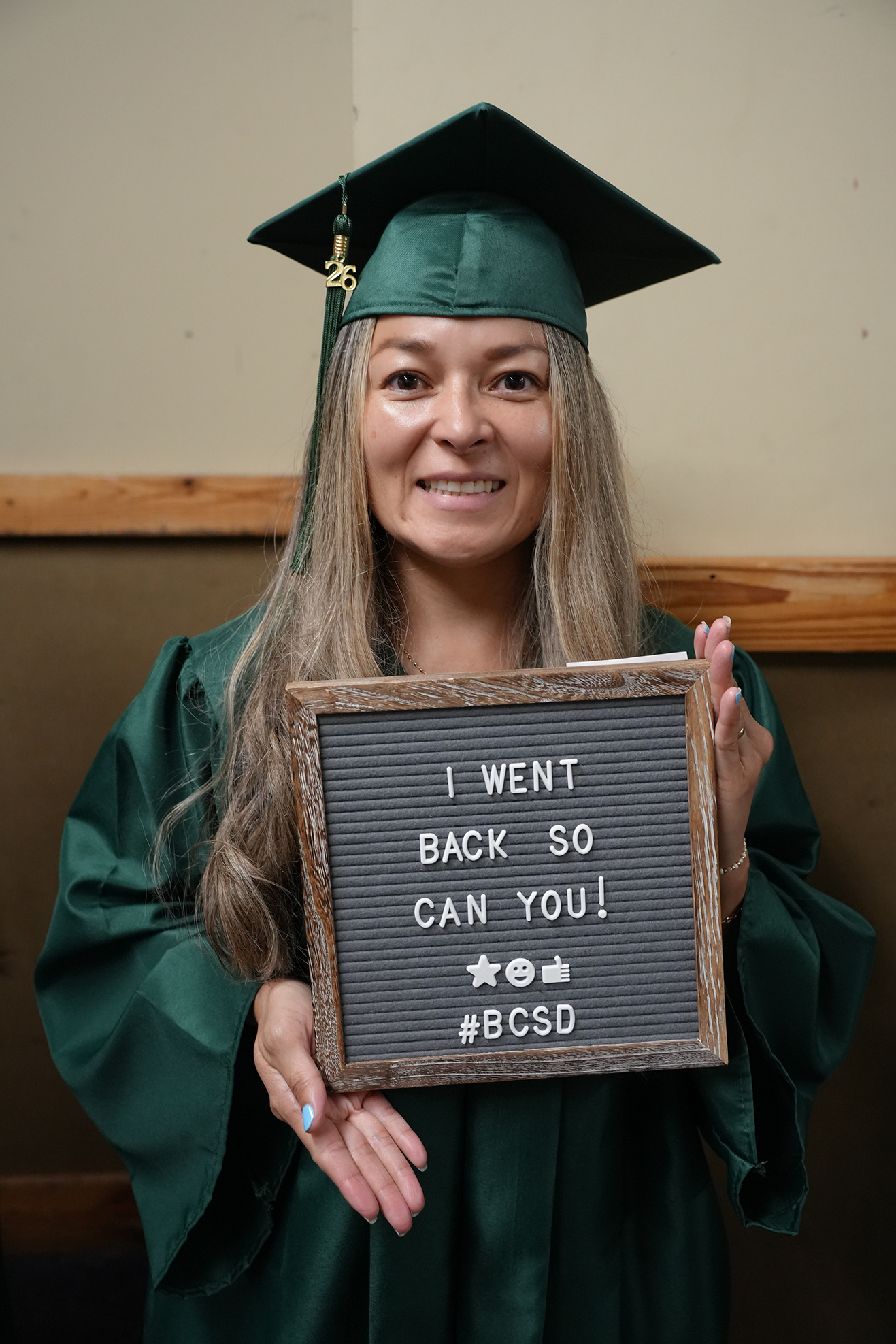 Woman in green graduation garb holding sign that says, "I WENT BACK SO YOU CAN YOU!"