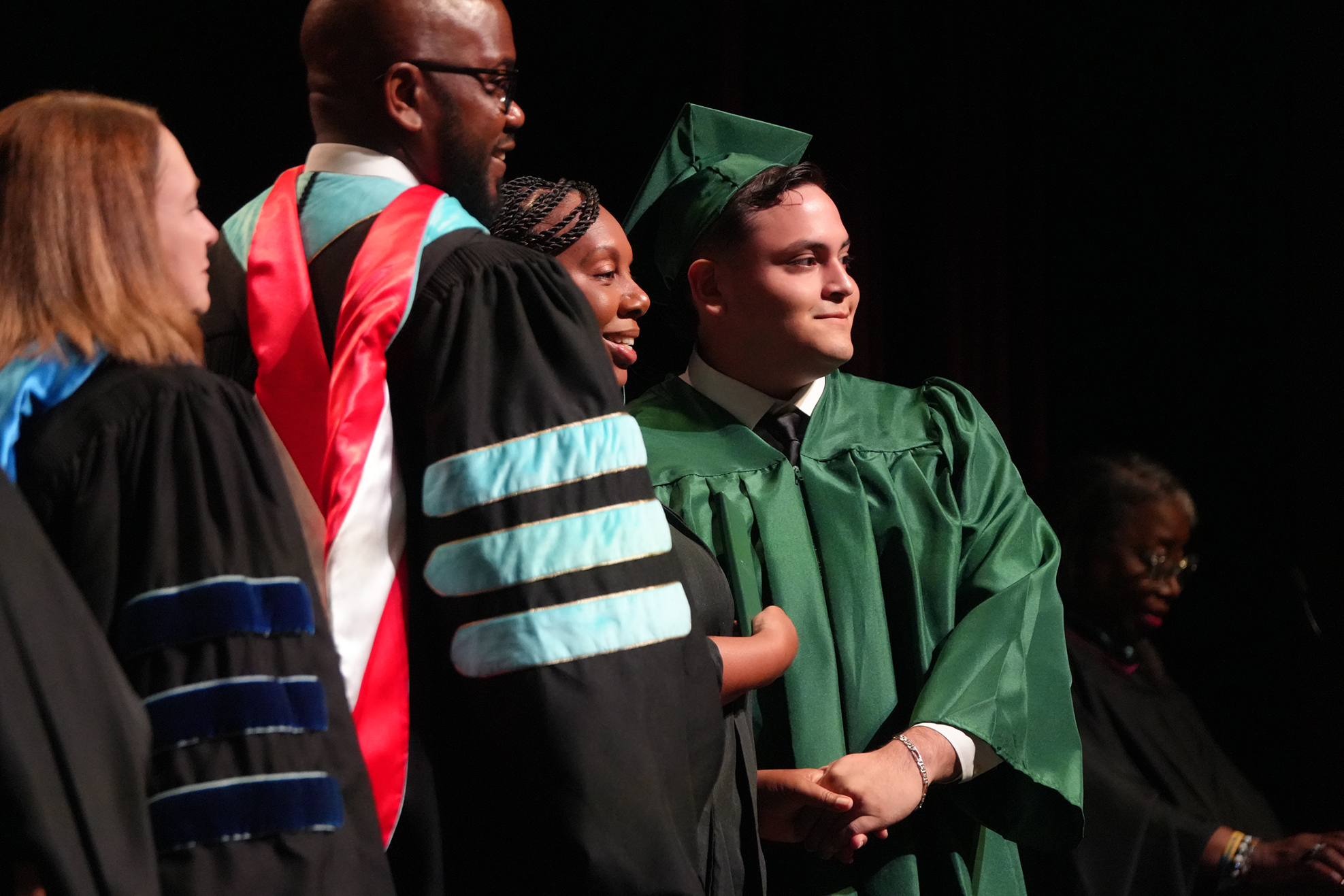 A group of people in academic regalia. A man in a green gown smiles as he shakes hands.