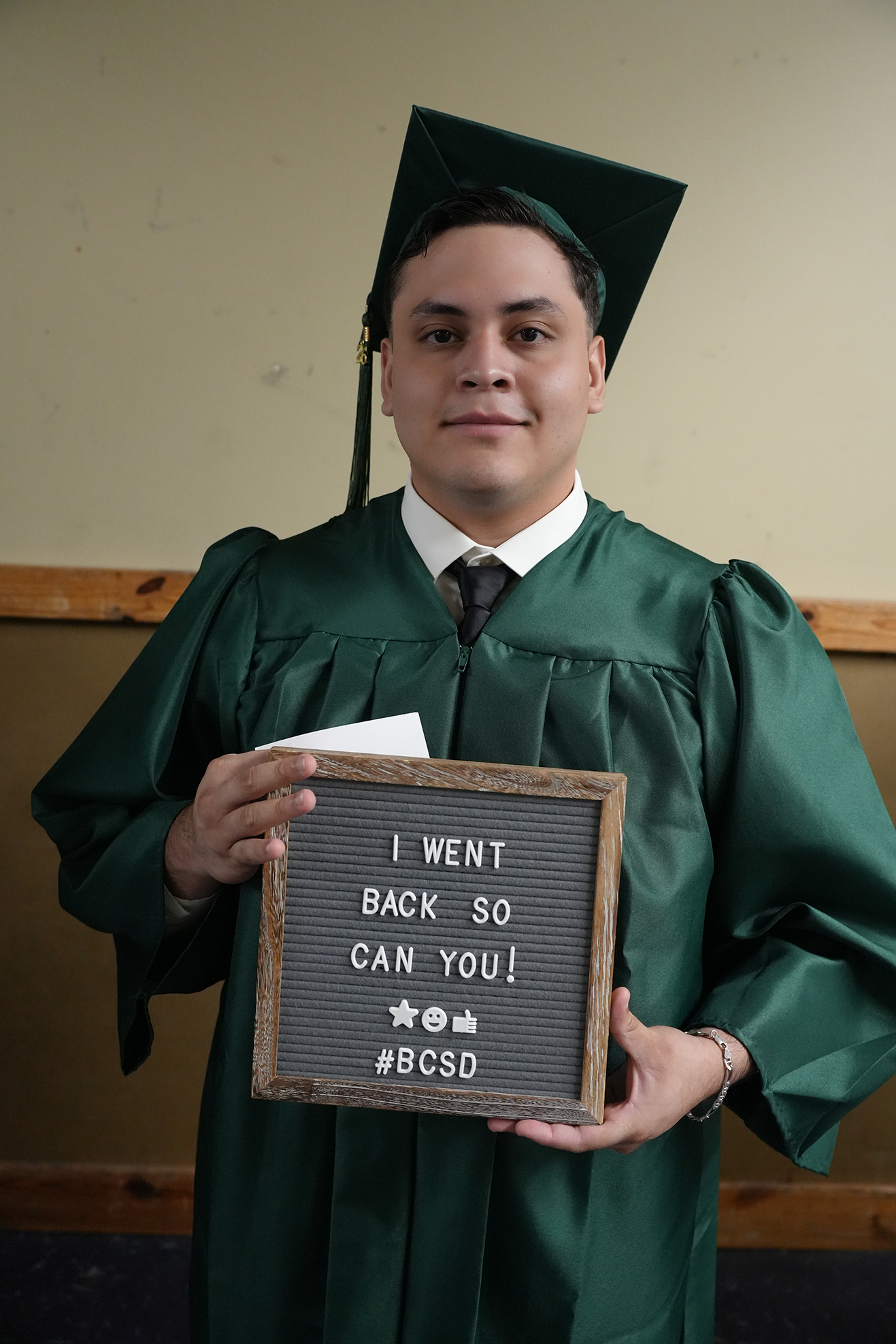 A man in a green graduation gown and cap holds a chalkboard that says, "I WENT BACK SO YOU CAN YOU! #BCSd."