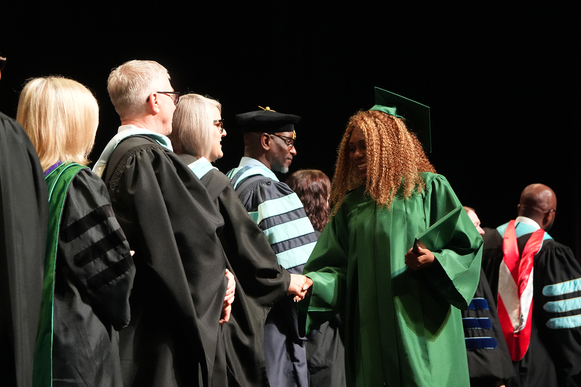 Several people in academic regalia stand on stage. A woman in a green gown and mortarboard faces the audience.