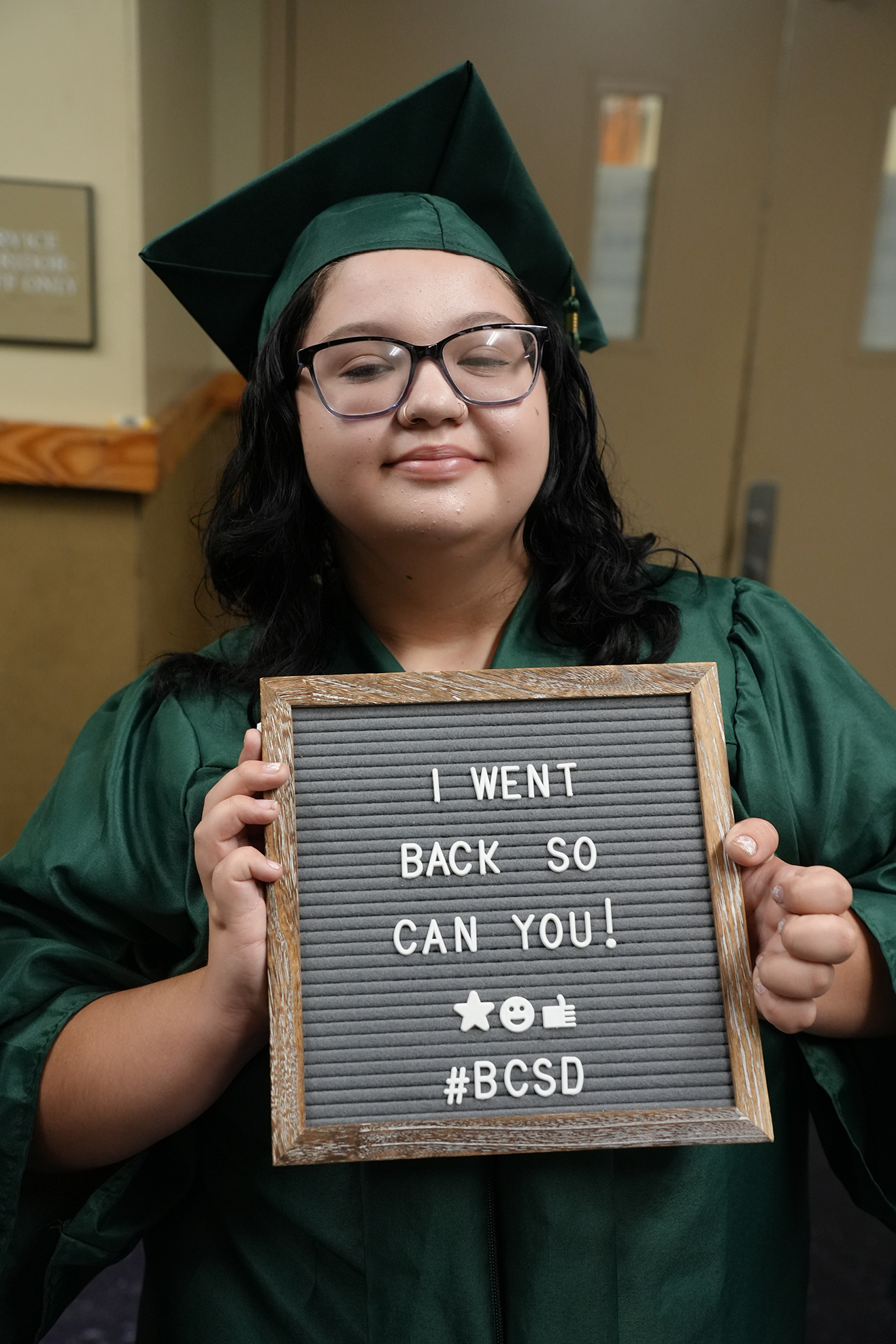 Person in graduation gown holding a sign that reads "I WENT BACK SO CAN YOU!" with a hashtag.