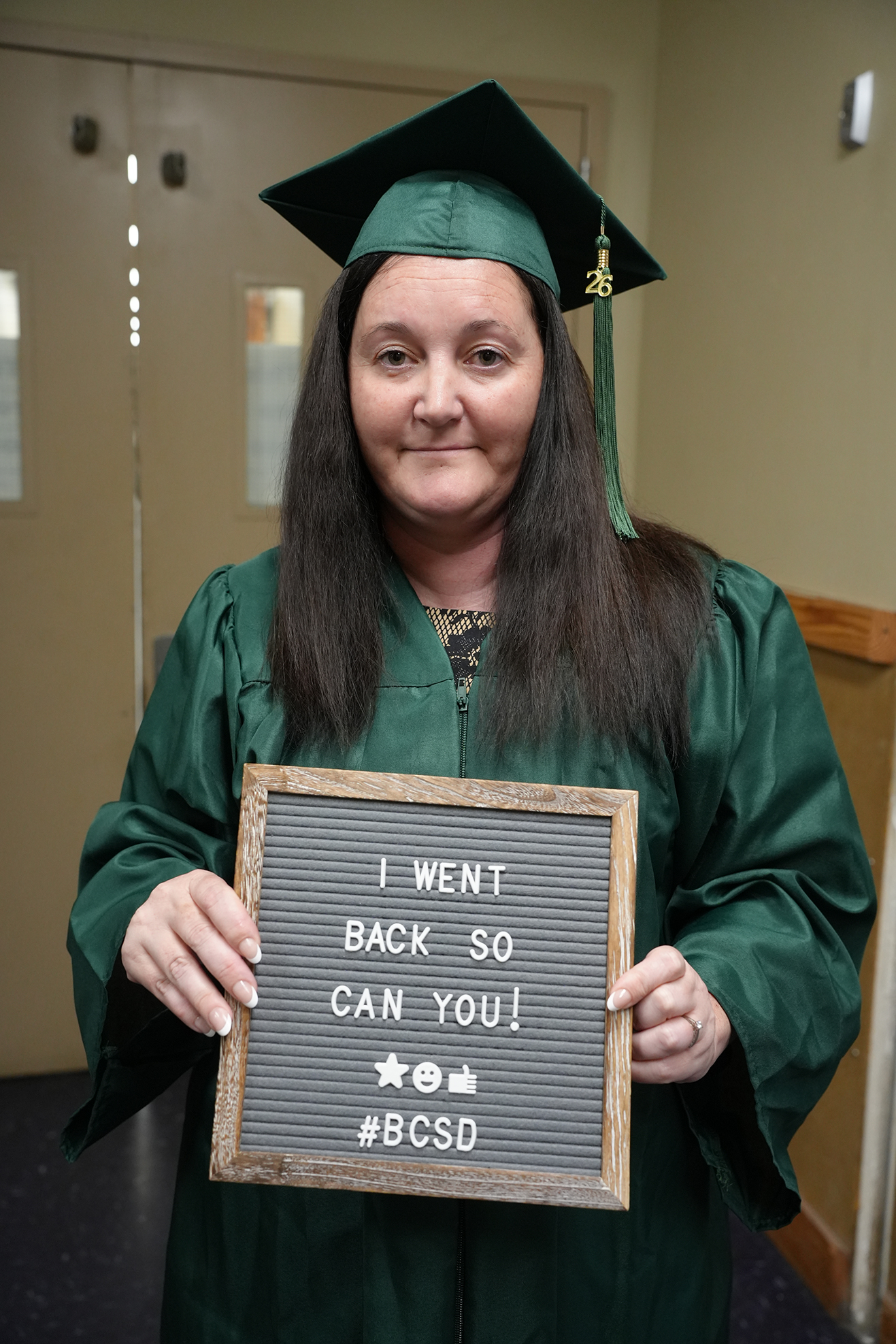 A woman in a graduation gown holds a sign with a message, "I WENT BACK SO YOU CAN TOO! #BCSD."