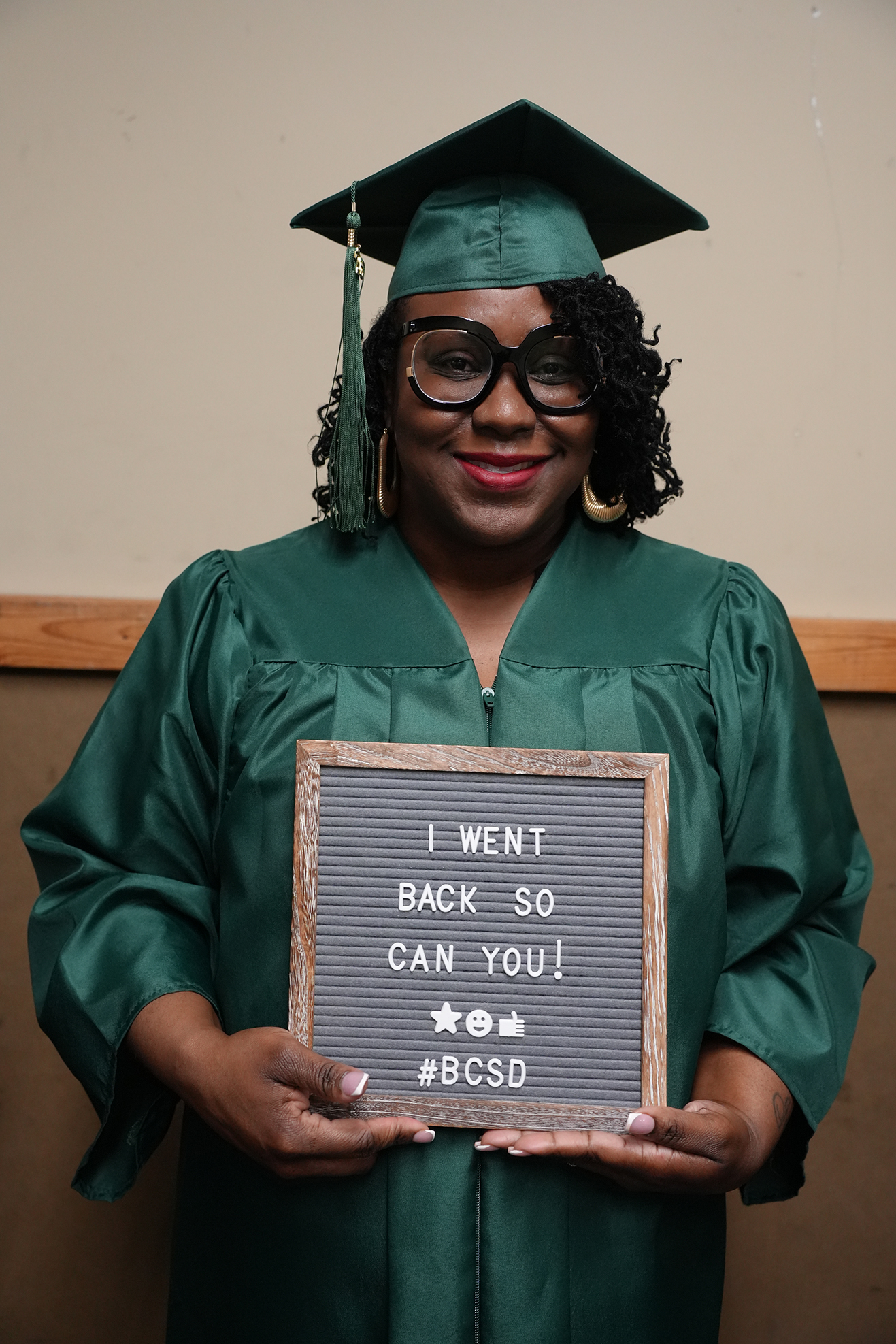 Person in a green graduation gown holding a sign that says "I WENT BACK SO CAN YOU!"