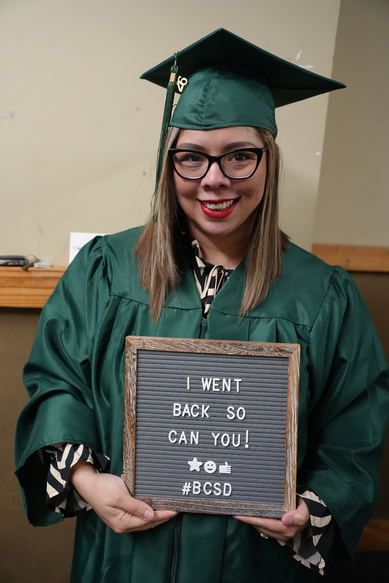 A woman in a green graduation gown holding a sign that says "I WENT BACK SO YOU CAN YOU!" with a backdrop of a beige wall.