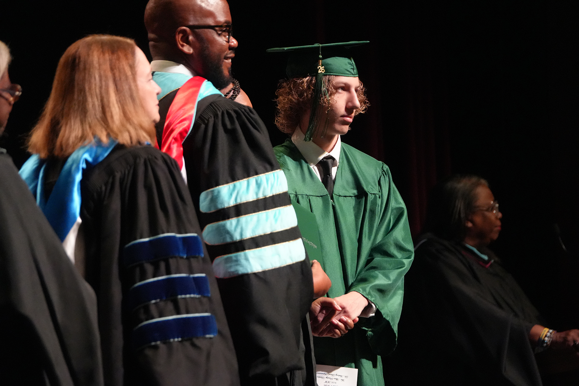Five adults in academic regalia stand on a stage, one holding the hands of another. One person wears a green gown.