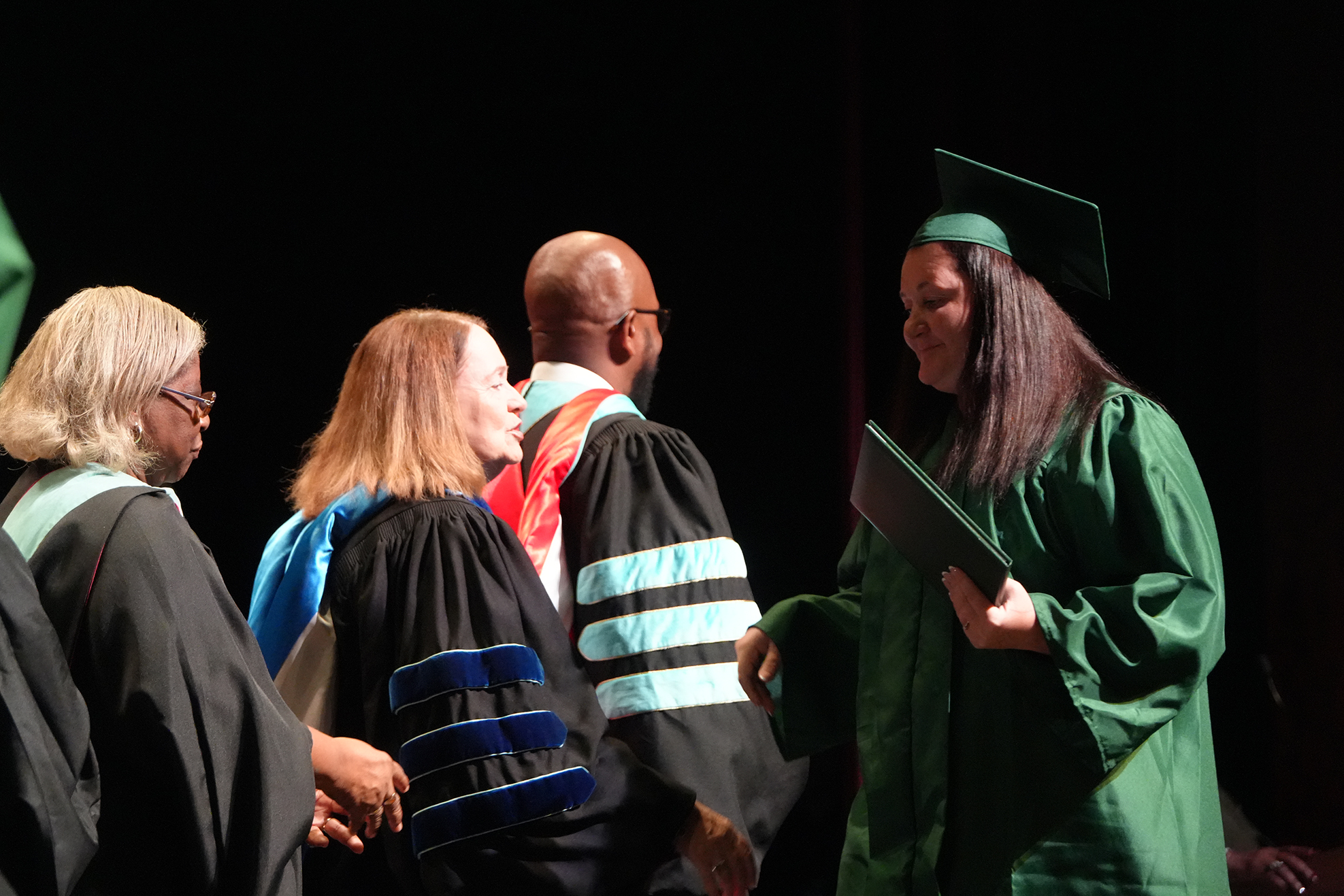 Five adults, each wearing academic regalia, stand on a stage. One holds a folder and looks toward another.