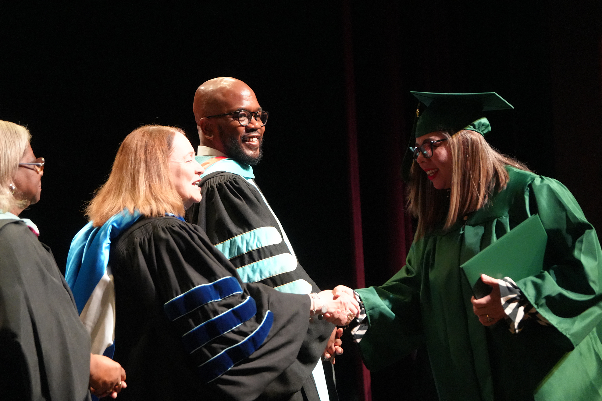 Three individuals in graduation gowns on a stage; a graduate shakes hands with a faculty member.