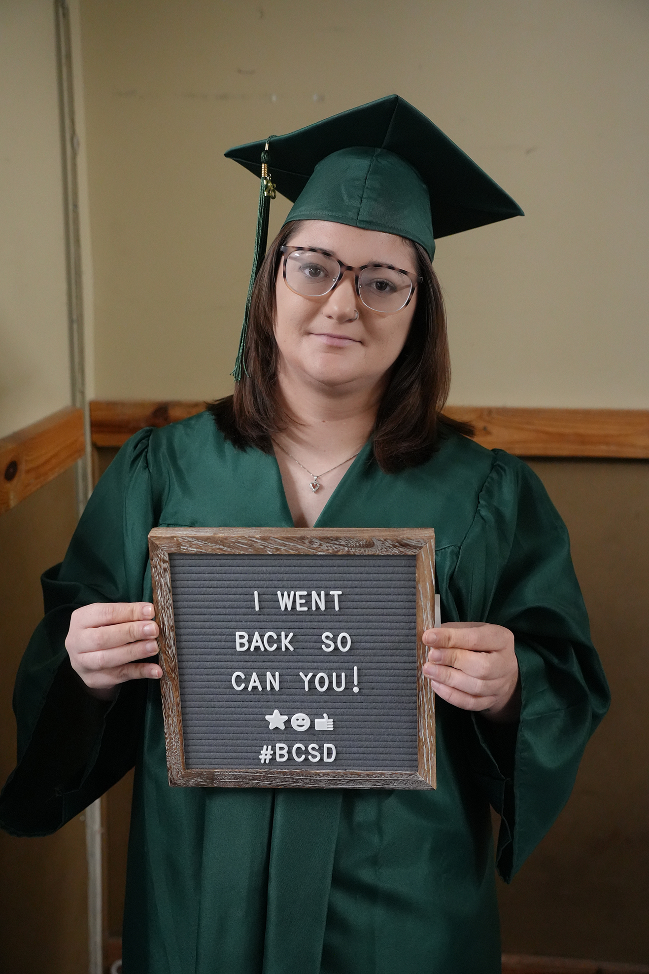 Person in green graduation gown holding a sign that says, "I WENT BACK SO CAN YOU!"