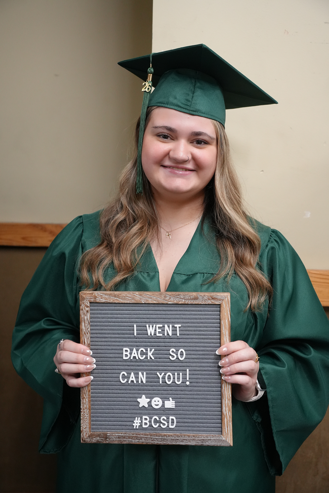 A woman in a green graduation gown and cap holds a sign that says "I WENT BACK SO YOU CAN YOU!" against a wall.