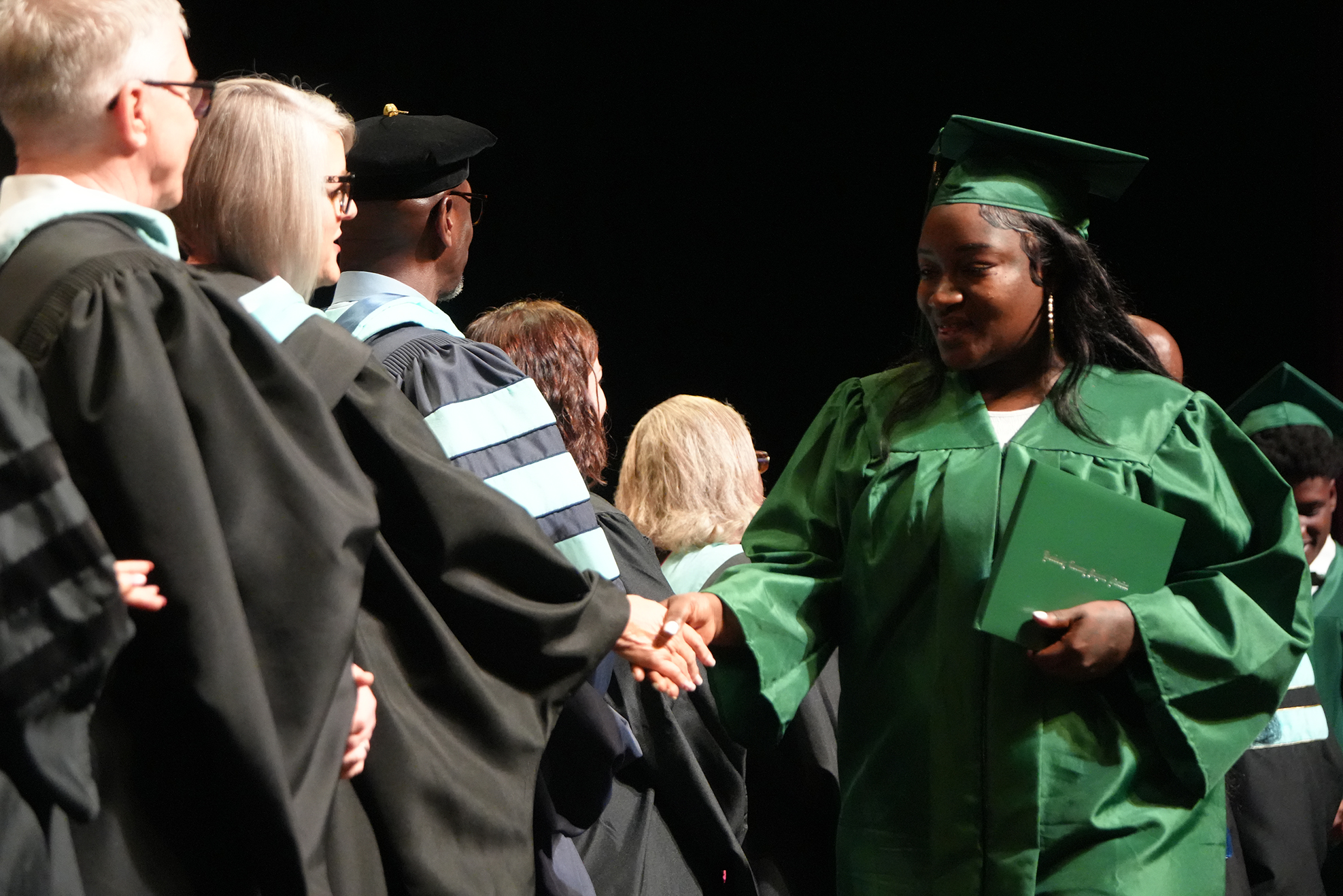 Graduates in green and black robes stand in line at an event; a graduate in green shakes hands with a graduate in black.