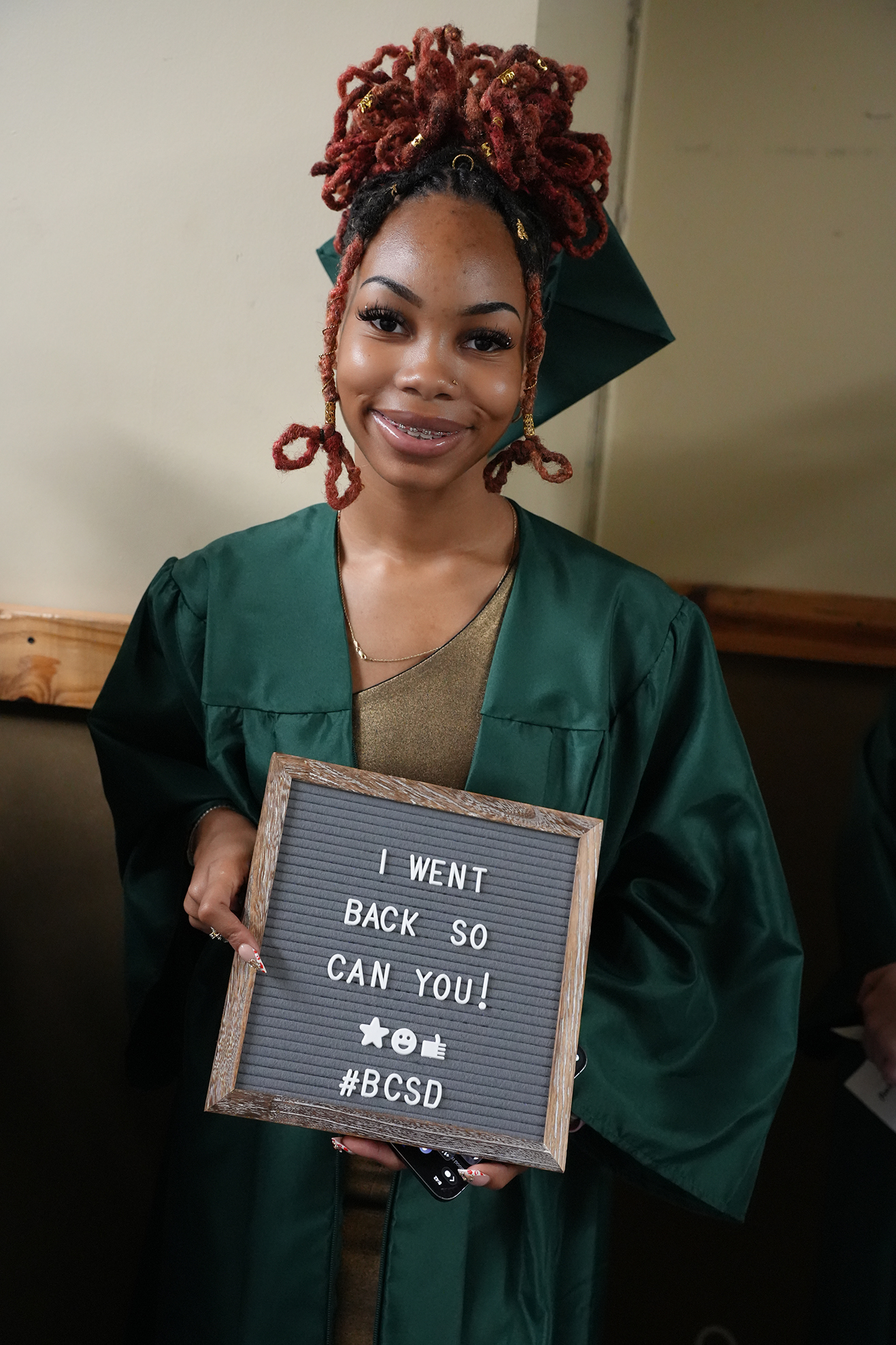 Woman in green graduation gown holds a sign reading "I went back so you can too! #BCCSD" and smiles.