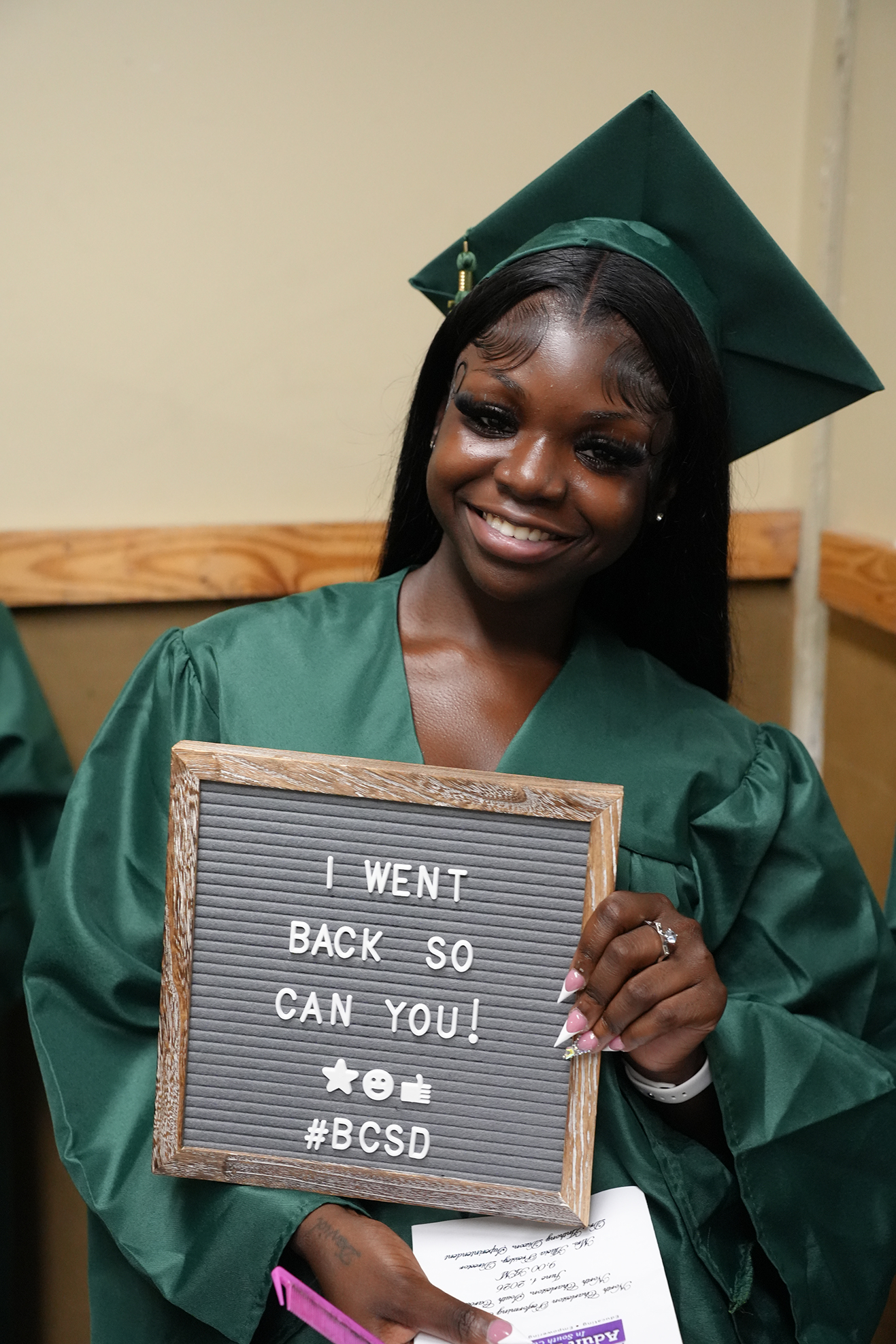 Woman in a green graduation gown holds a sign that reads, "I WENT BACK SO CAN YOU! #BCSD."