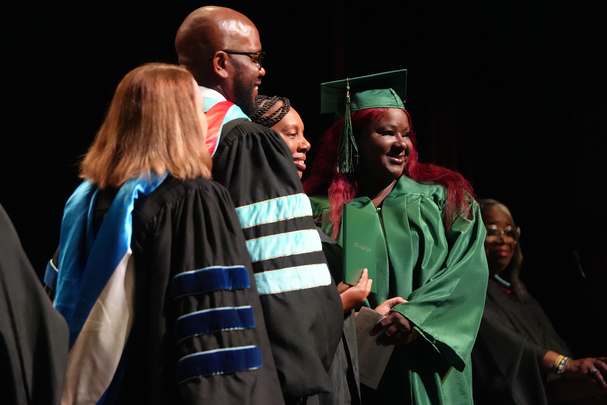 A woman in green graduation garb smiles as she stands among other graduates in black robes.